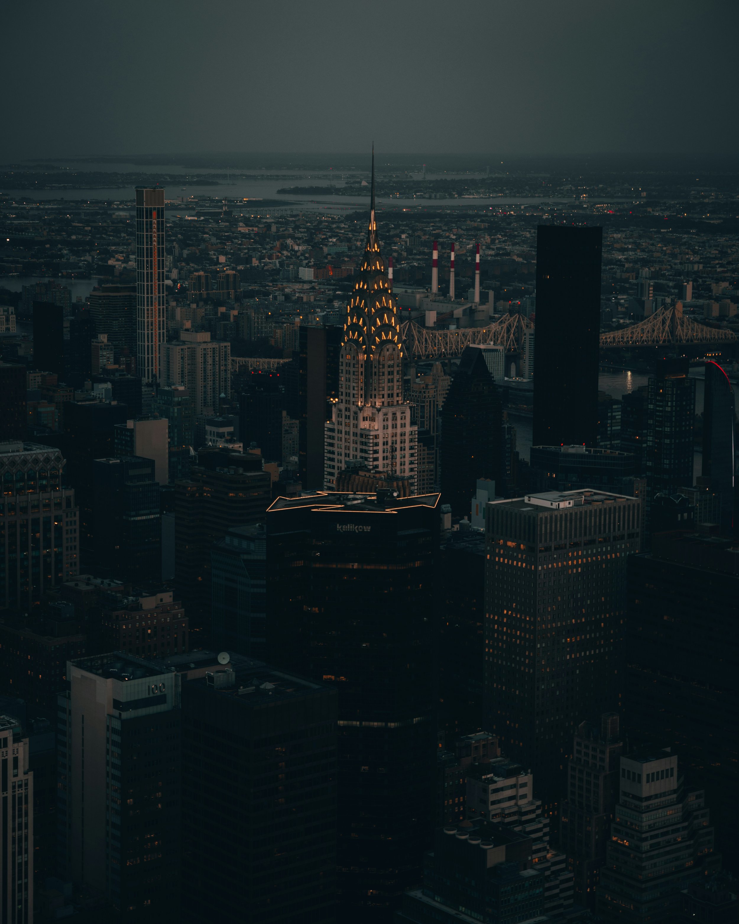 Nighttime aerial view of Manhattan with the Chrysler Building illuminated, surrounded by tall skyscrapers and bridges in New York City.