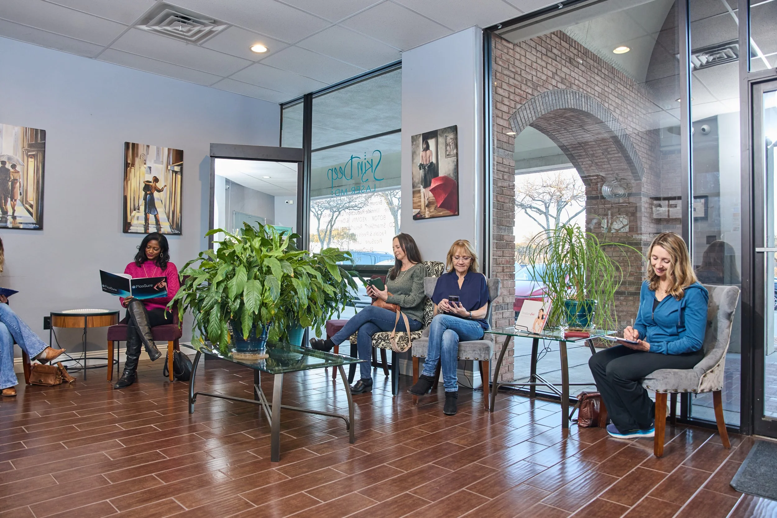 People sitting in a waiting area of an office or clinic with large windows, paintings on the wall, and potted plants.