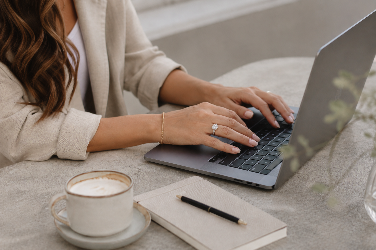 Person typing on a laptop at a table, with a notebook and pen nearby.