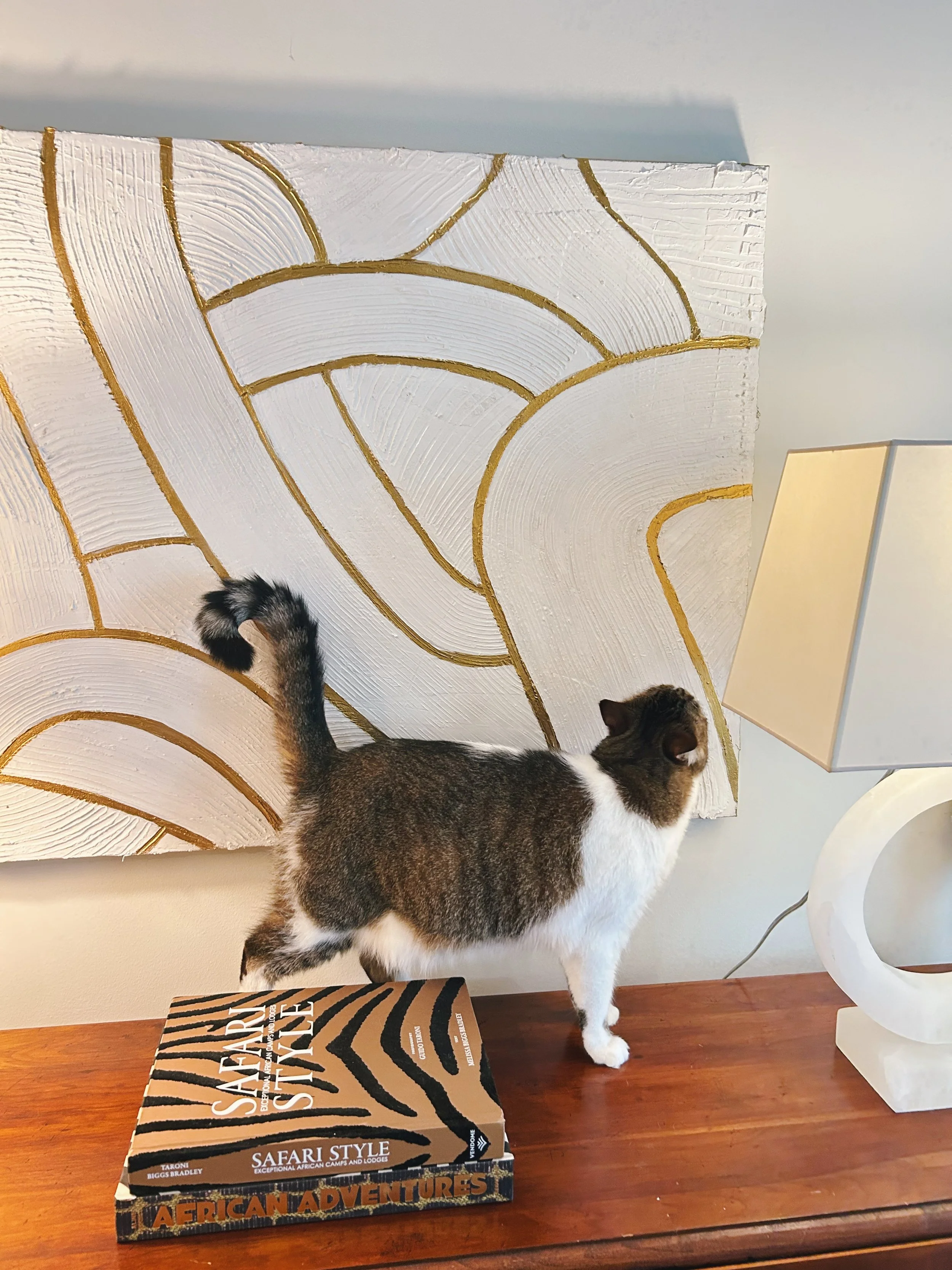 A tabby and white cat standing on a wooden table, with its head near a white, modern, sculptural table lamp and a decorative wall art piece with gold lines on a textured white background. There is a box labeled 'Safari Style' on the table.