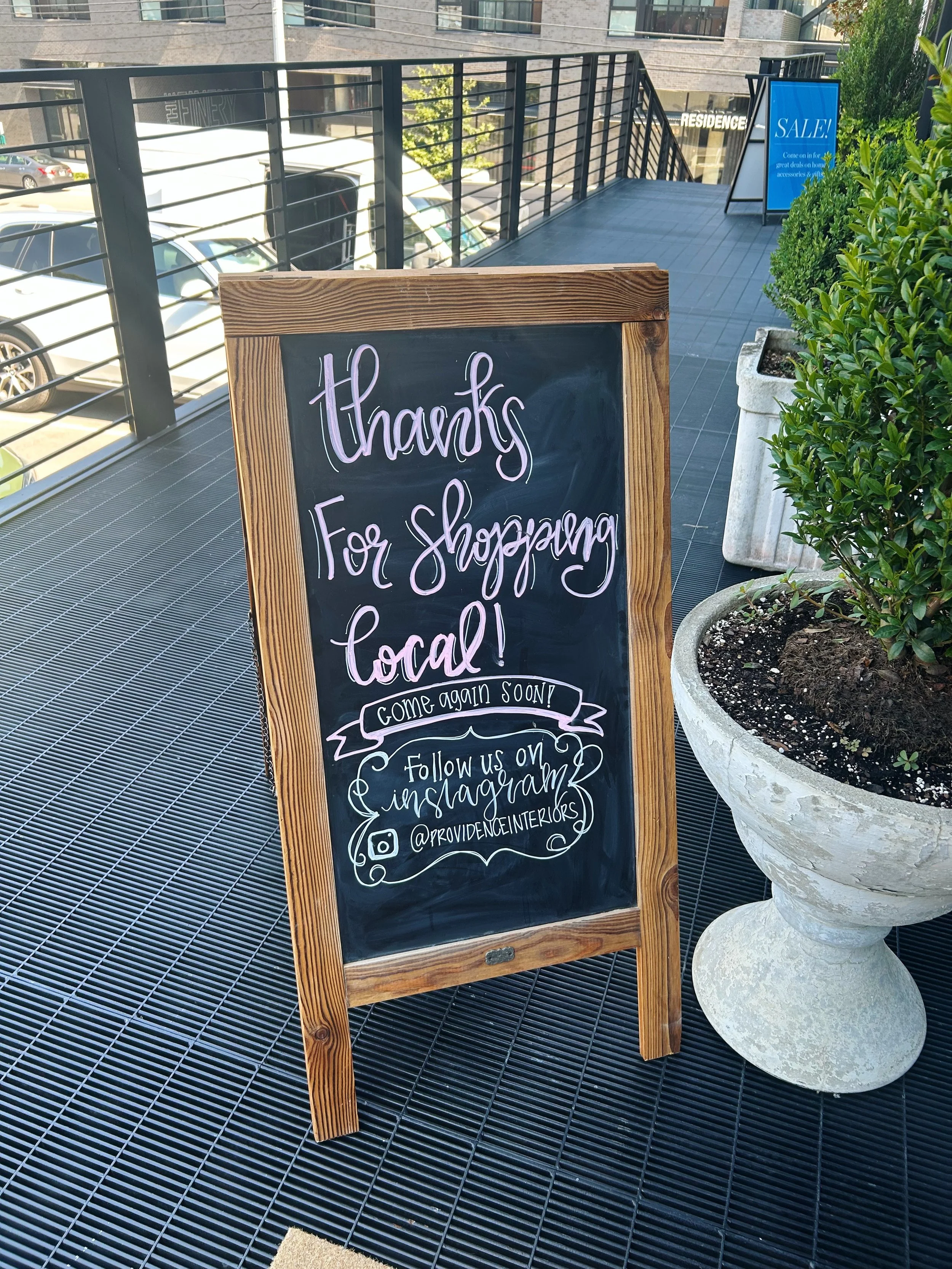 A chalkboard sign thanking customers for shopping locally, encouraging them to come back soon and follow on Instagram, placed next to a large planter on an outdoor balcony with parking and buildings in the background.