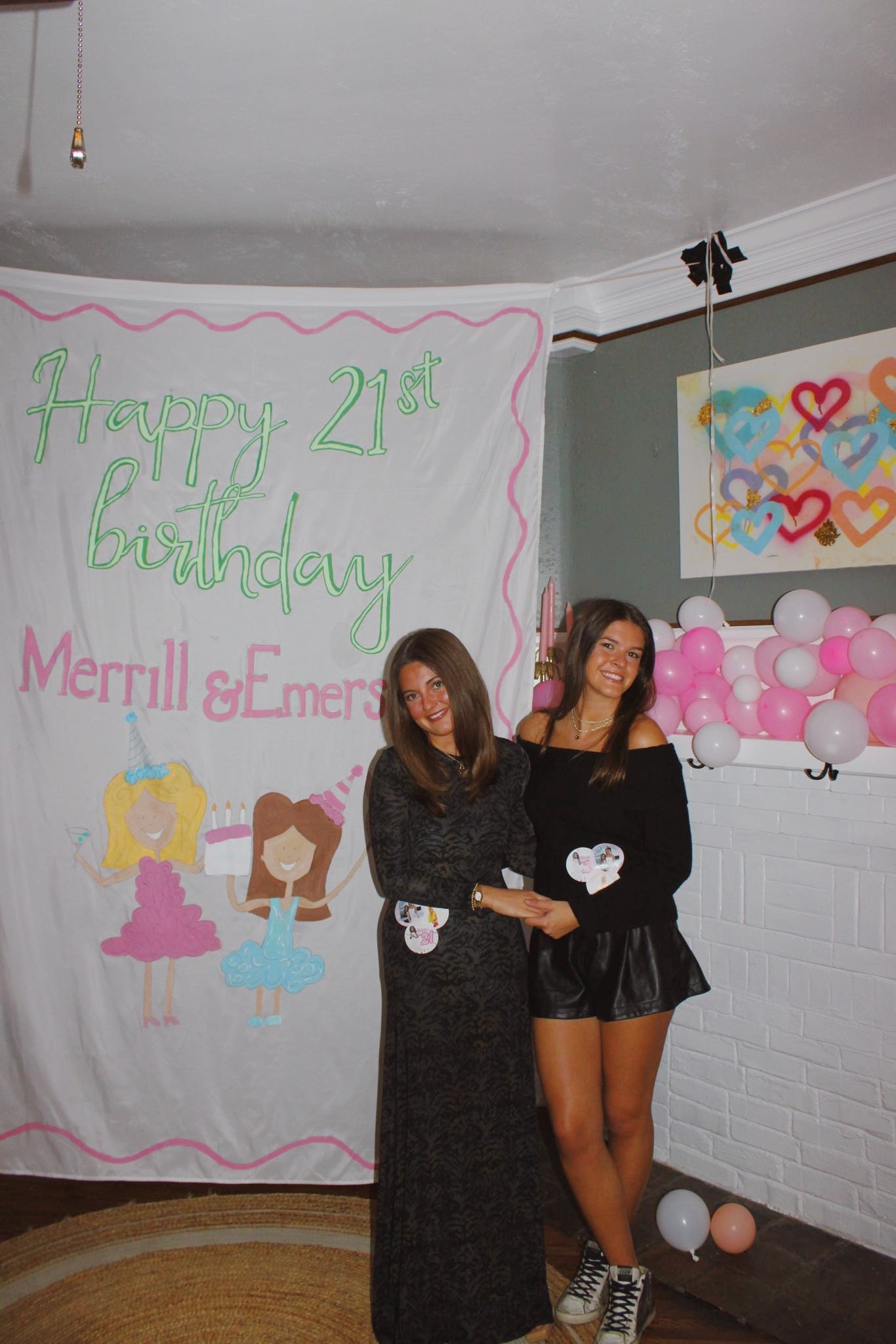 Two women standing in front of a birthday banner that says 'Happy 21st Birthday Merrill & Emers'. The women are smiling and holding hands, with pink and white balloons decorating the background.