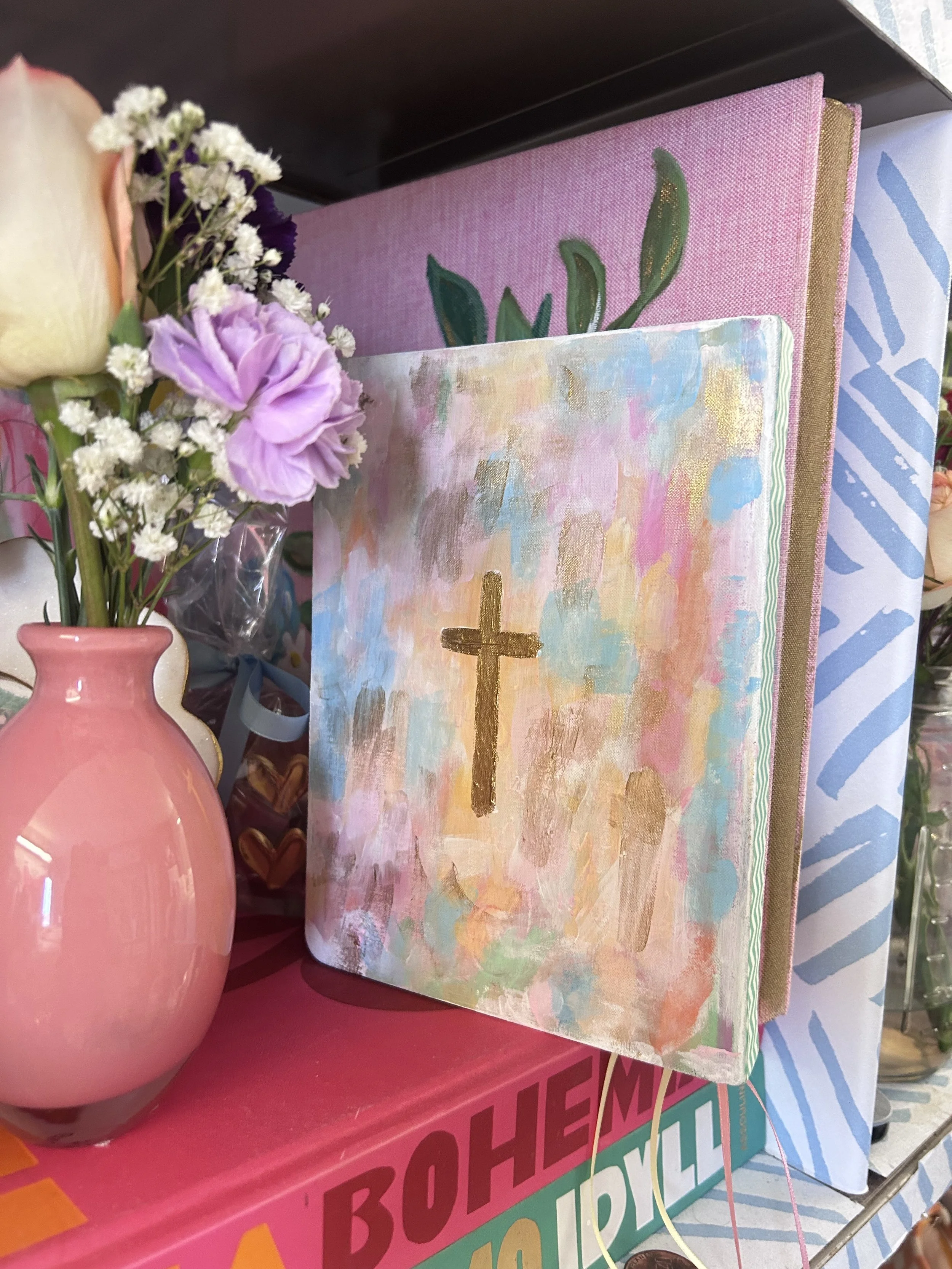 A pink vase with white and purple flowers, beside a stack of colorful notebooks, with a small painting of a cross on a pastel background leaning against the notebooks.