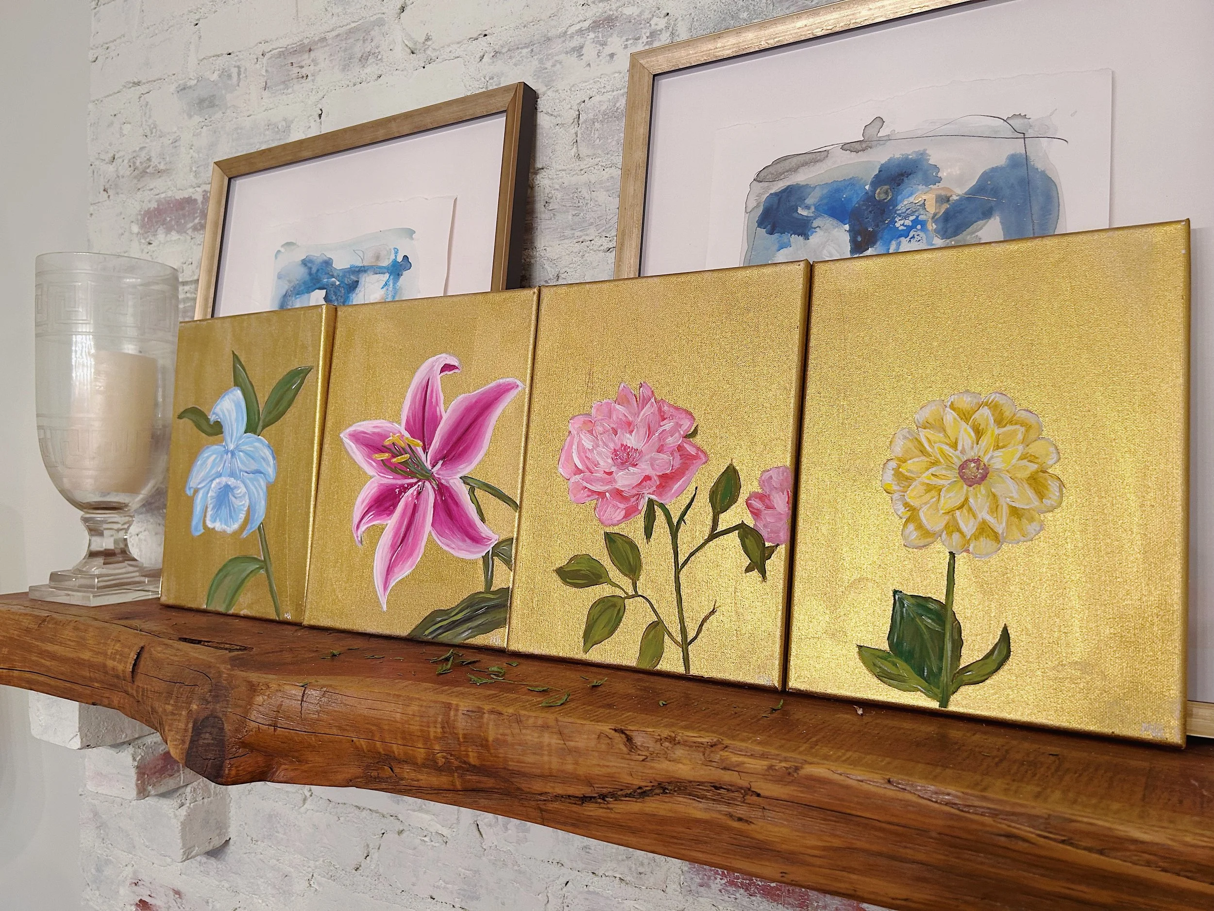 Four floral paintings with pink, blue, and yellow flowers on gold backgrounds, placed on a wooden shelf against a white brick wall, with two framed abstract watercolor paintings in a wooden frame above.