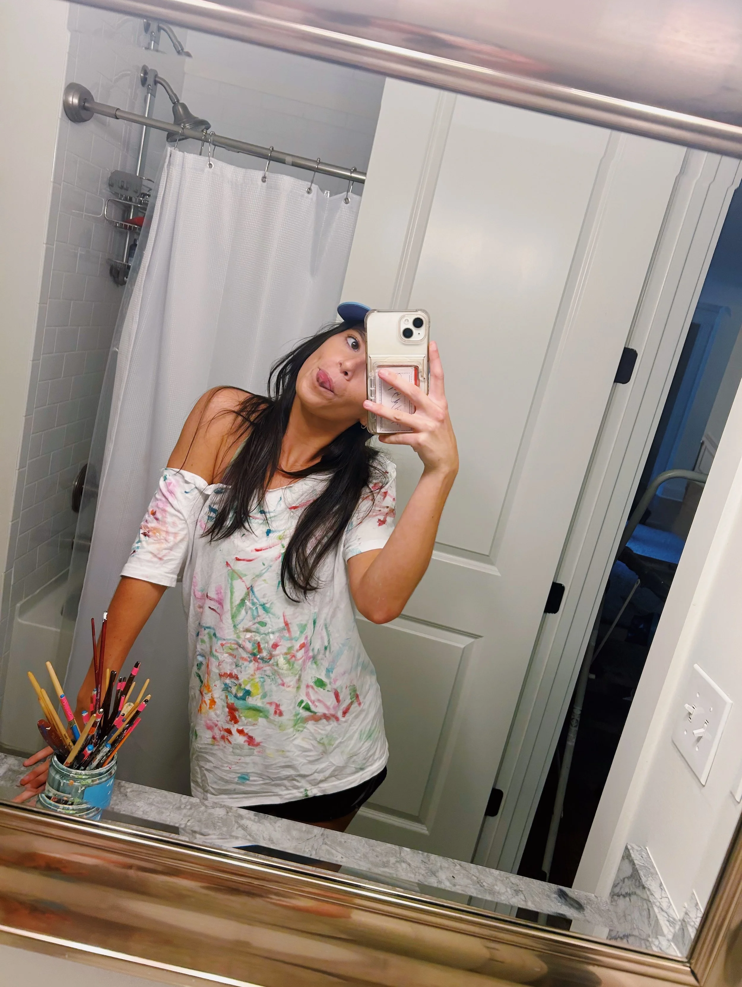 Woman taking a mirror selfie in a bathroom, wearing a colorful, paint-stained t-shirt and black shorts, with a shower and closet in the background.