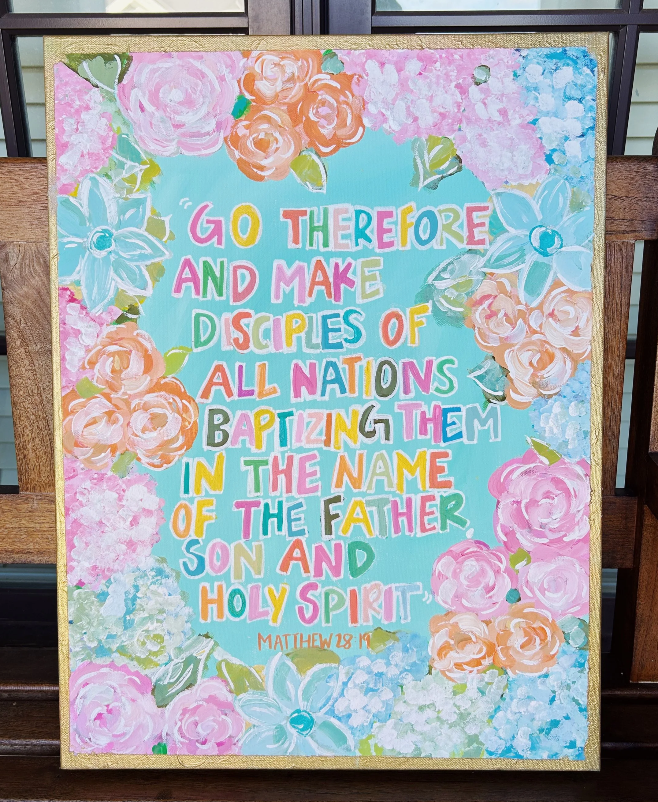 Colorful painted sign with floral border featuring pink, peach, and blue flowers. Text from Matthew 28:19, "Go therefore and make disciples of all nations, baptizing them in the name of the Father, Son, and Holy Spirit."