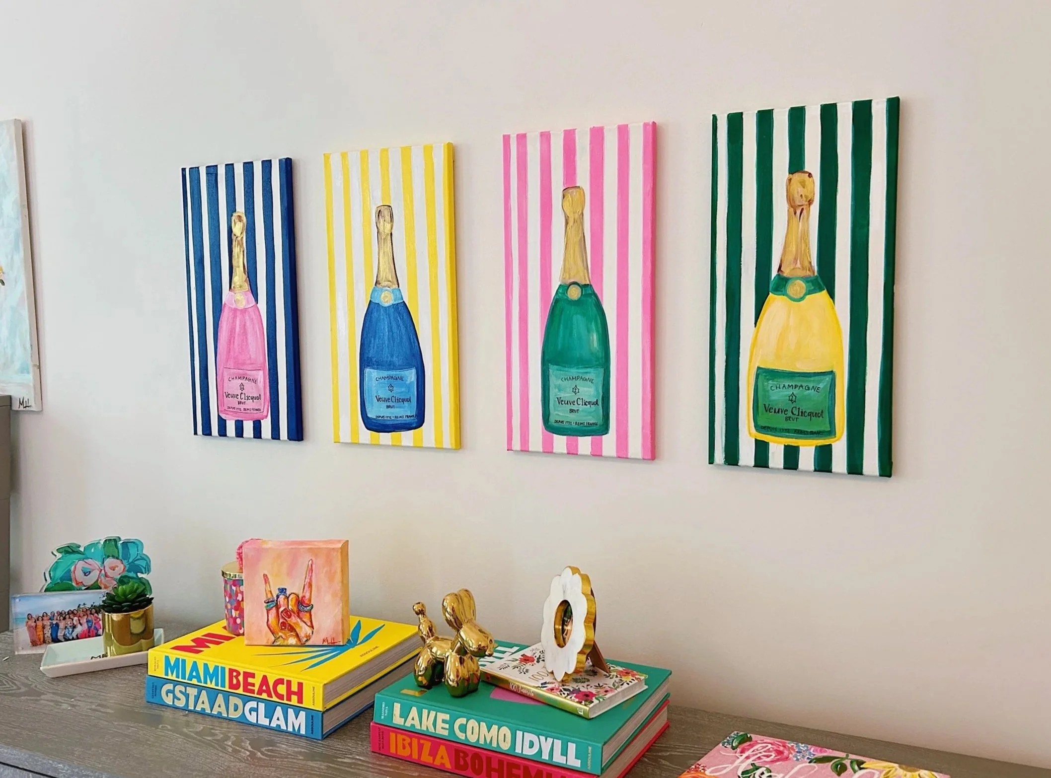 Colorful artwork of champagne bottles on a white wall, with bottles in pink, blue, green, and yellow backgrounds on striped canvases. Below, a table holds books, a small plant, gold figurines, and decorative items.