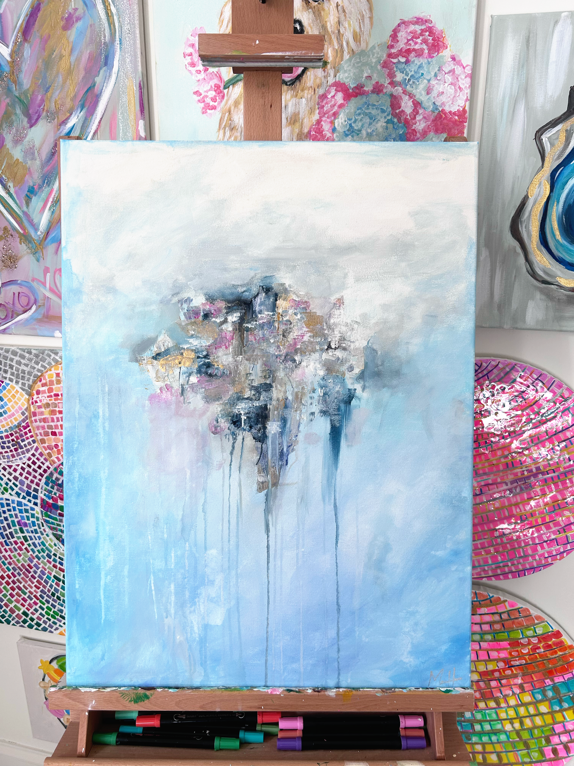 An abstract painting of a boat, dock, and water scene in soft pastel colors including blue, pink, and white, on an easel in an art studio surrounded by other colorful artworks.