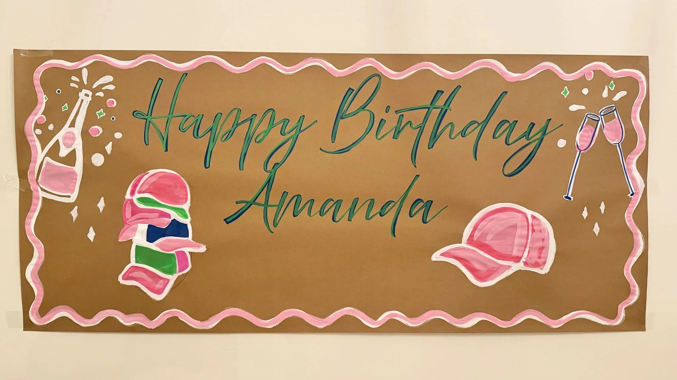 Decorative birthday banner with the message 'Happy Birthday Amanda' surrounded by illustrations of a champagne bottle popping, two clinking champagne glasses, and a pink baseball cap, all outlined in pink and with festive accents.