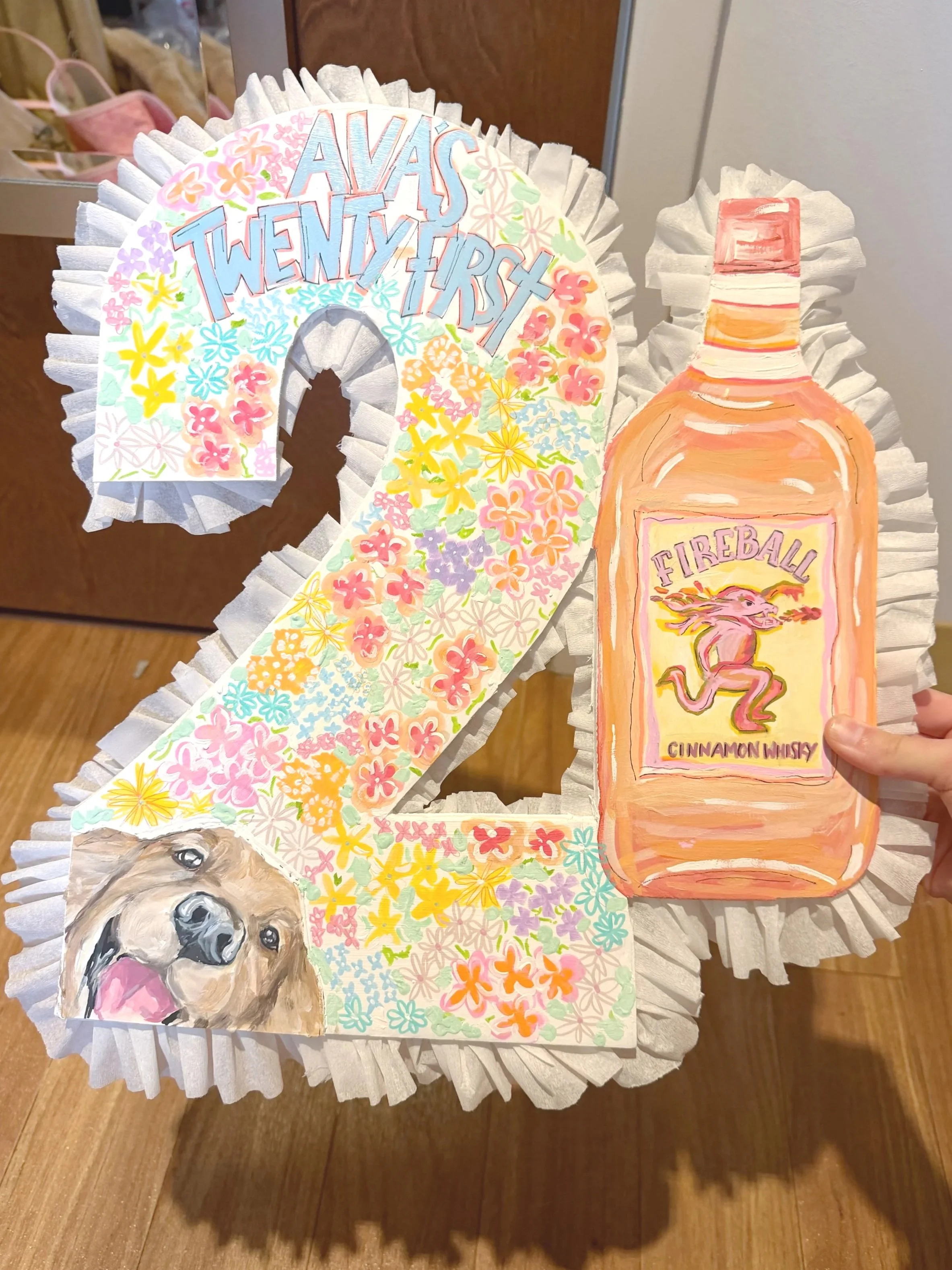 Handmade decorative piece featuring a large floral wreath with the phrase 'AVAS TWENTY FIRST' written on it, a painted bottle labeled 'Fireball Cinnamon Whisky', and a portrait of a golden retriever puppy surrounded by colorful flowers, all on a whit