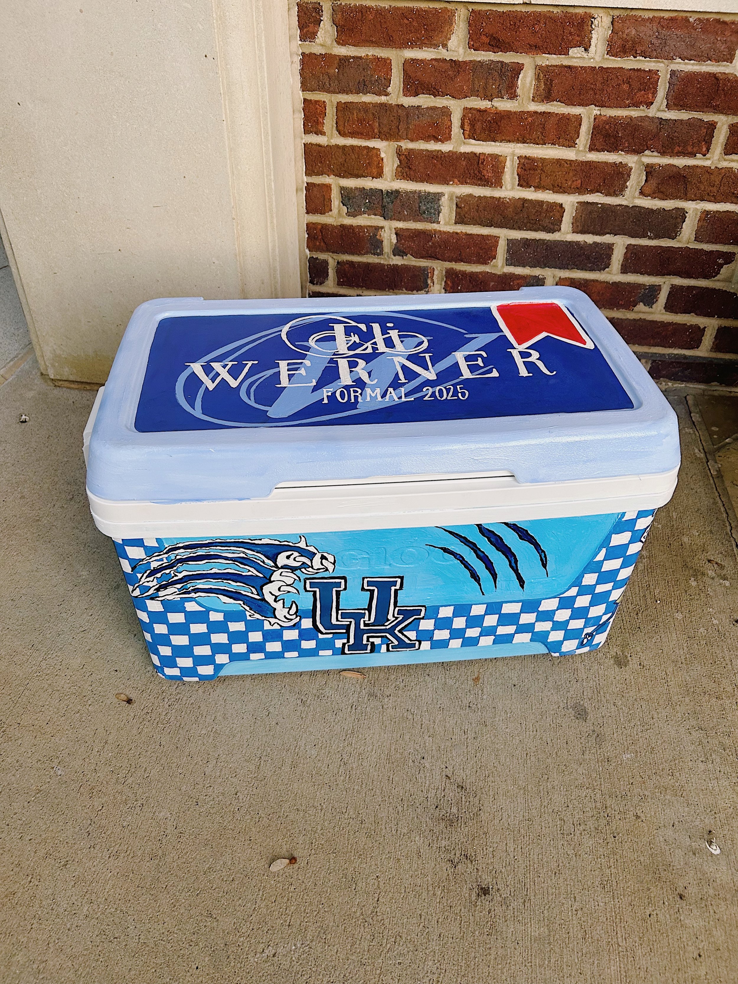 A painted cooler featuring the Kentucky Wildcats logo, with blue and white checkered pattern and claw marks, against a brick wall and concrete surface.