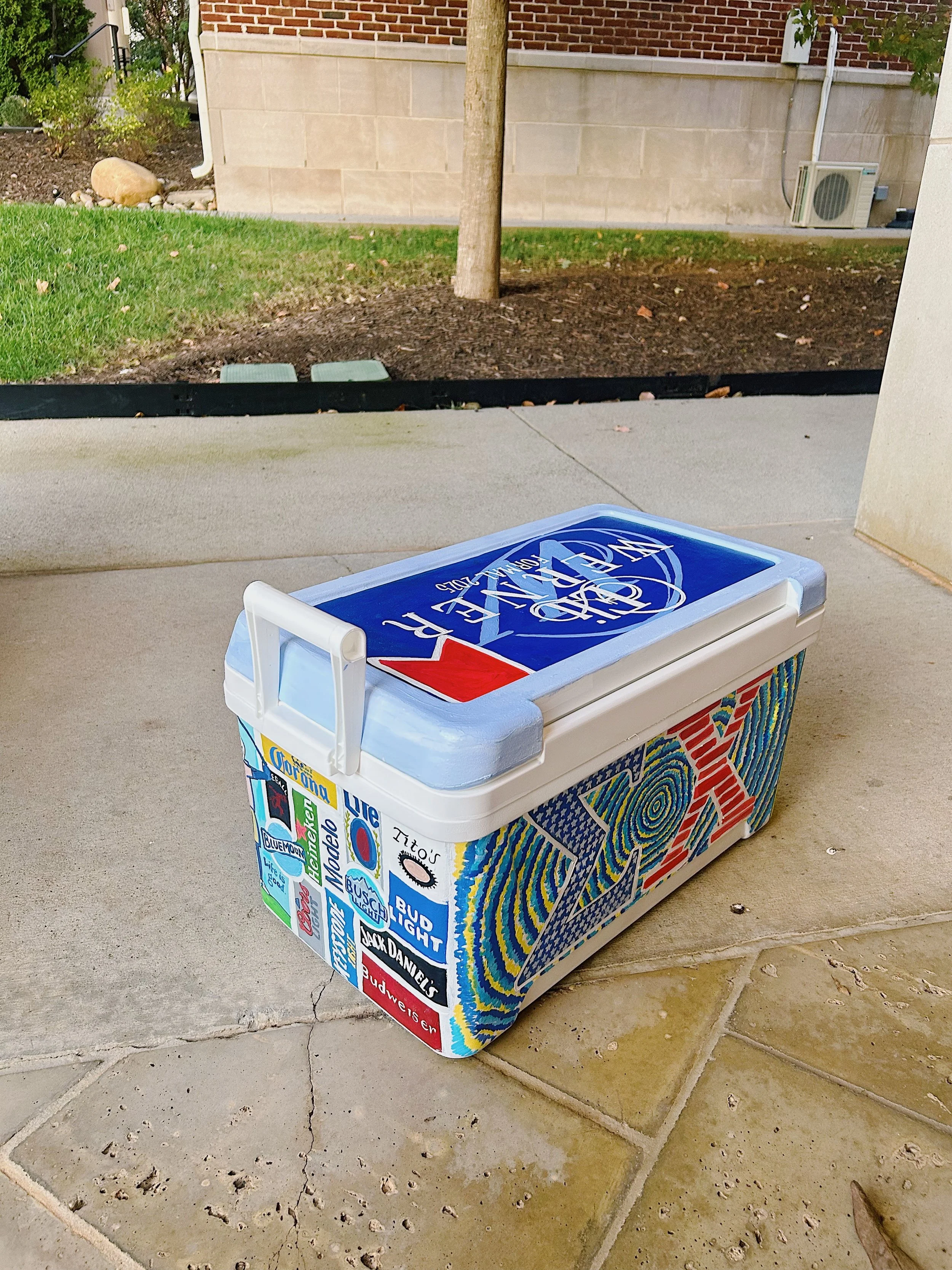Decorated cooler with colorful designs and logos, sitting on the ground outside on a sidewalk near a grassy area with a tree.
