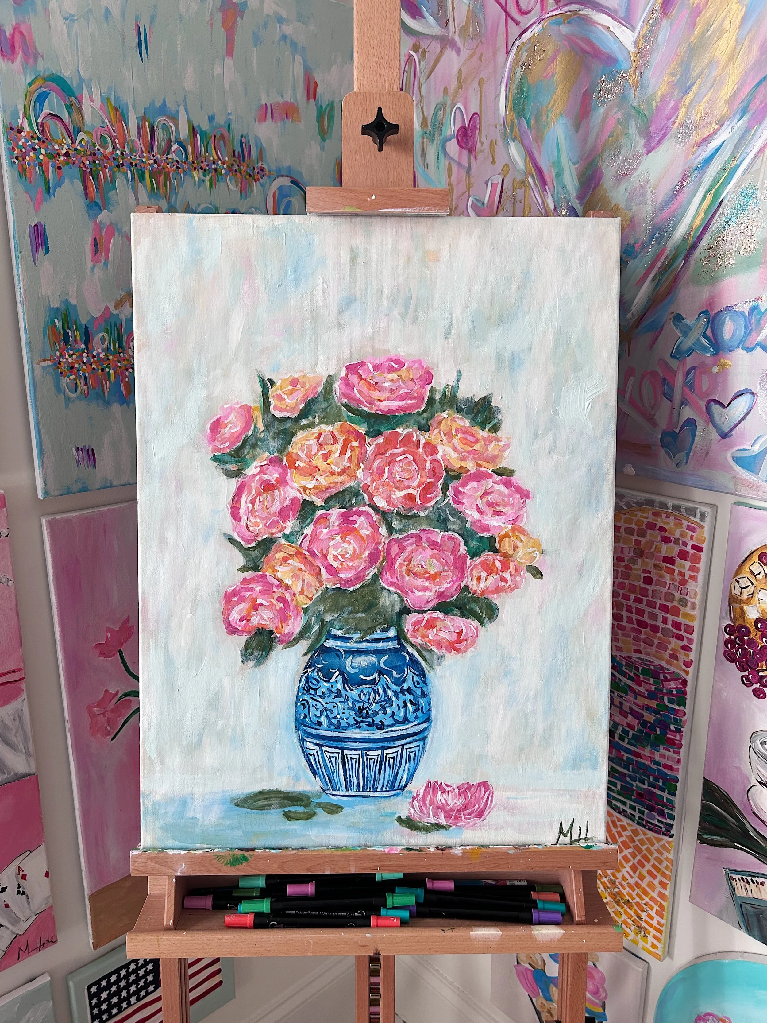 Painting of pink and peach flowers in a blue vase, positioned on an easel in an art studio with colorful murals on the walls.