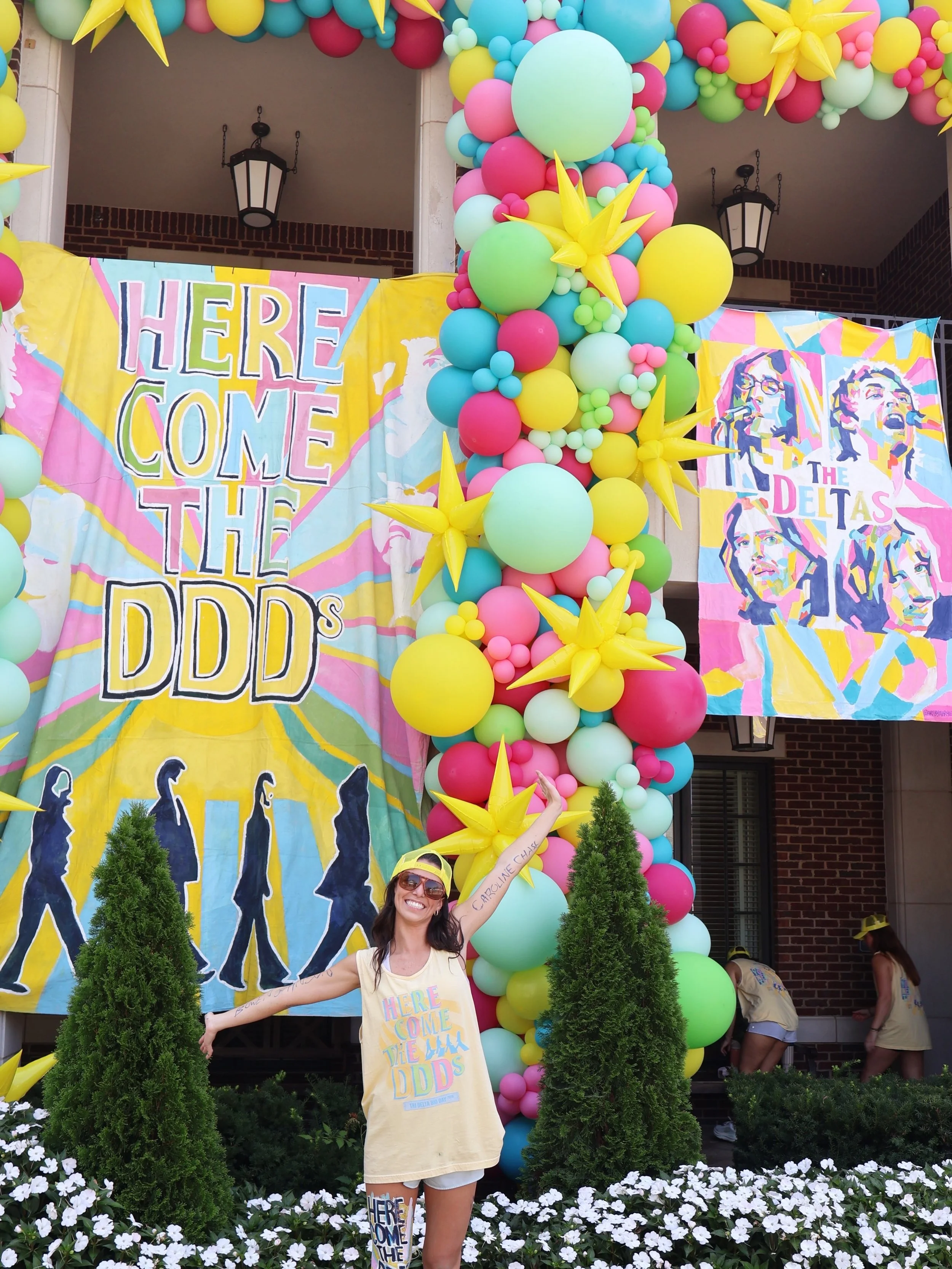 Woman smiling at a pride parade with balloons and banners, wearing a cream T-shirt with rainbow-colored text, standing in front of colorful balloon arch and floral decorations.