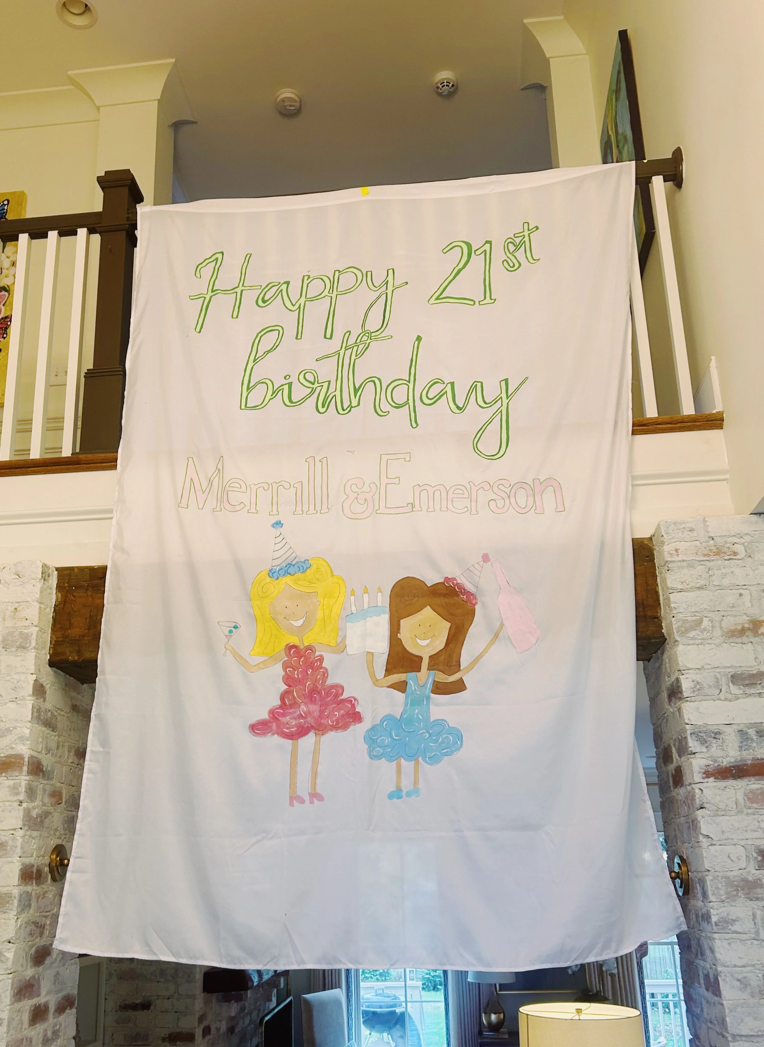 A birthday banner hanging inside a house with a brick wall and a fireplace. The banner has colorful hand-drawn illustrations of two girls celebrating, each holding a party item, with candles and a cake. The text reads, "Happy 21st birthday Merrill & 