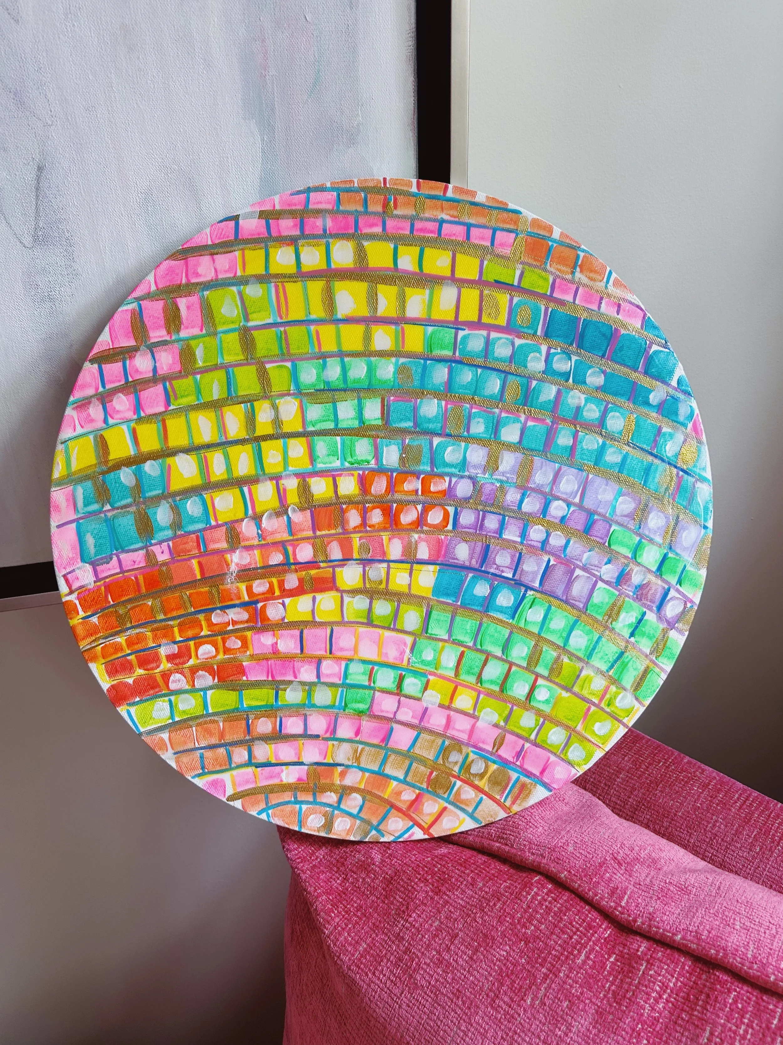 Colorful abstract circular artwork with a pattern of curved lines and small squares, placed on a pink textured surface.