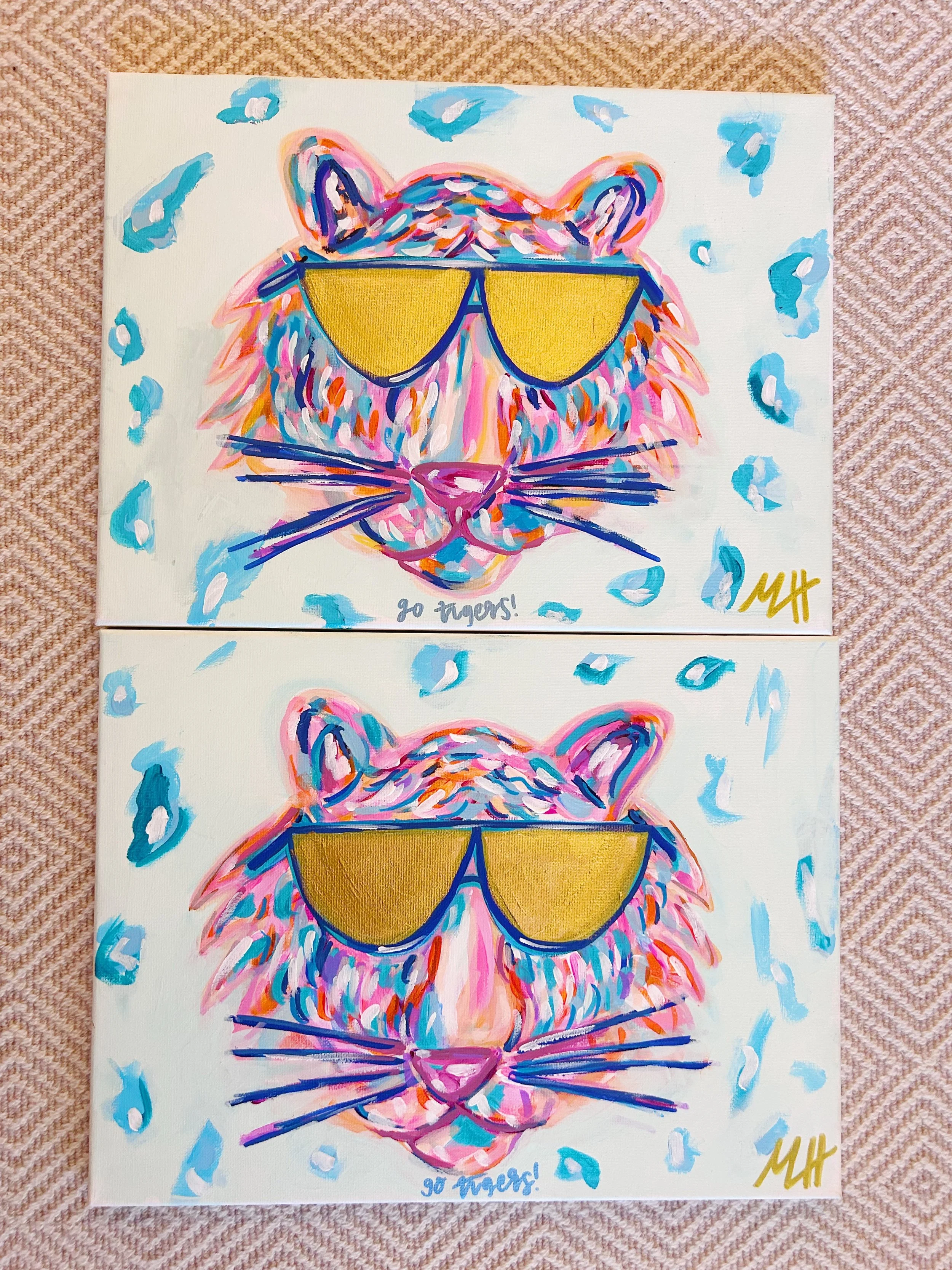Two colorful paintings of a lion wearing yellow sunglasses, surrounded by blue and pink abstract shapes, signed with initials 'MHK'. One painting has the words 'Go Tigers!' written underneath.