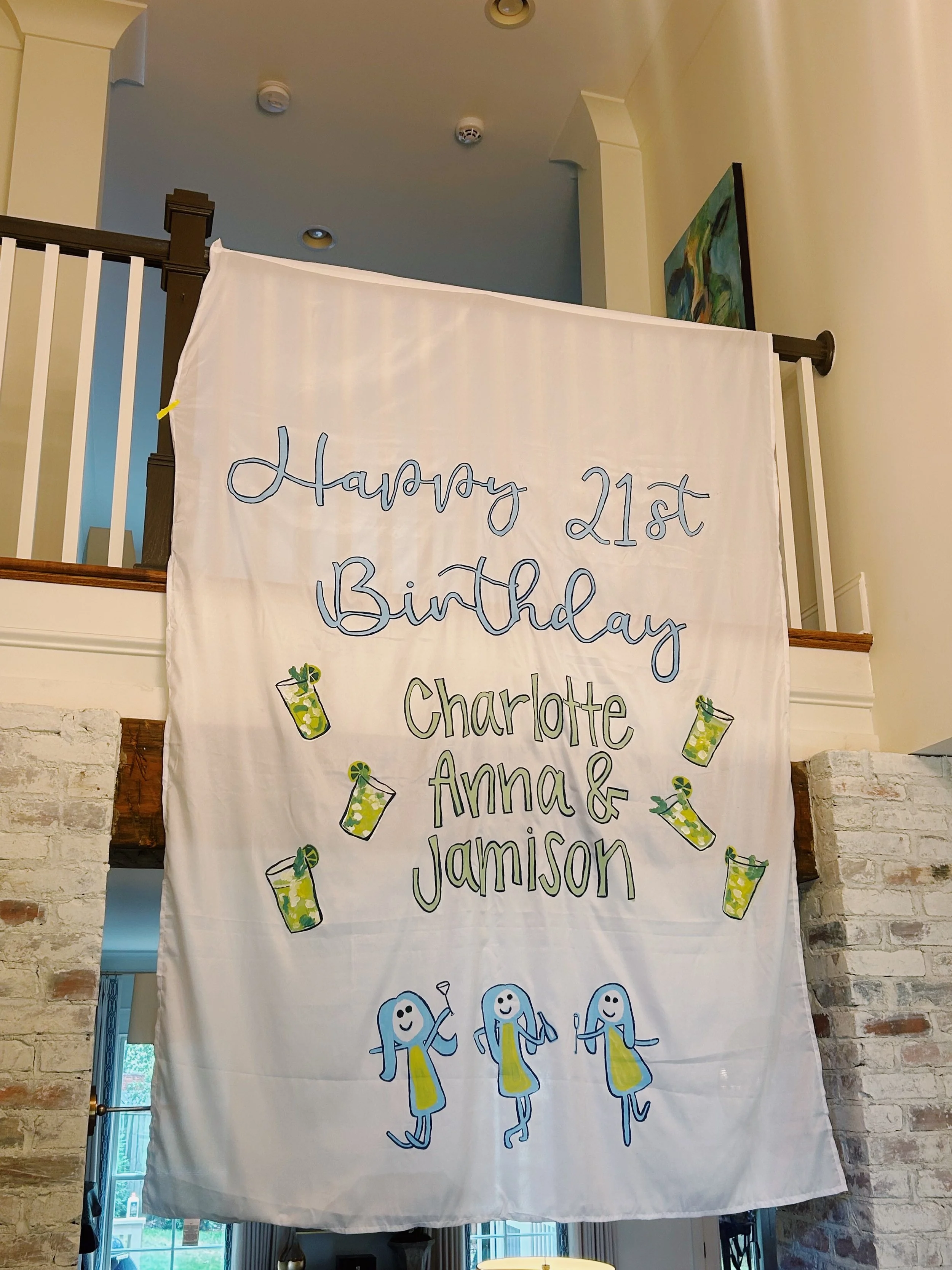 A large white banner with handwritten text that reads 'Happy 21st Birthday Charlotte, Anna & Jamison' and colorful illustrations of drinks and dancing figures.