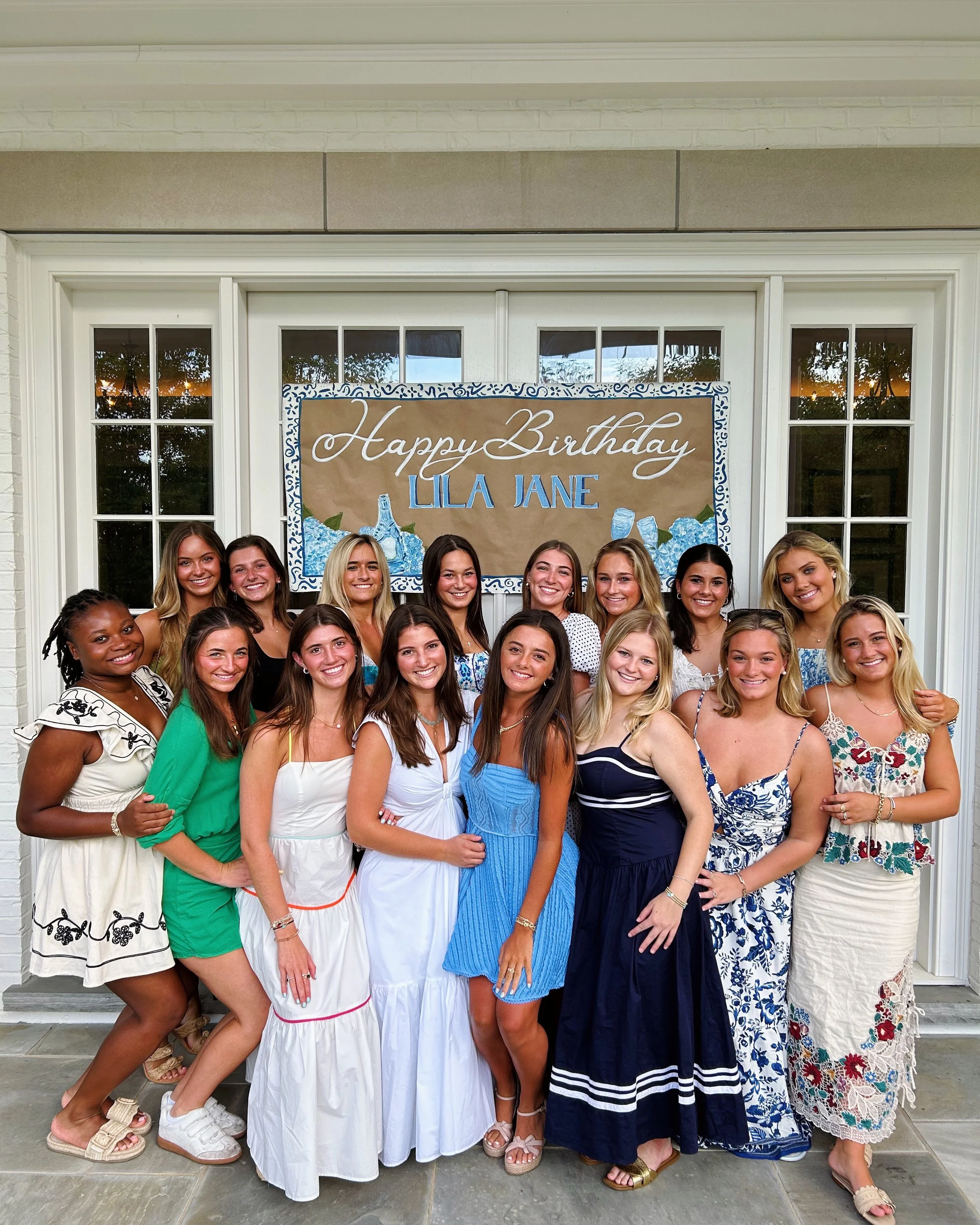 A group of young women dressed in colorful summer dresses, posing together at a birthday celebration in front of a large banner that says "Happy Birthday Lila Jane."