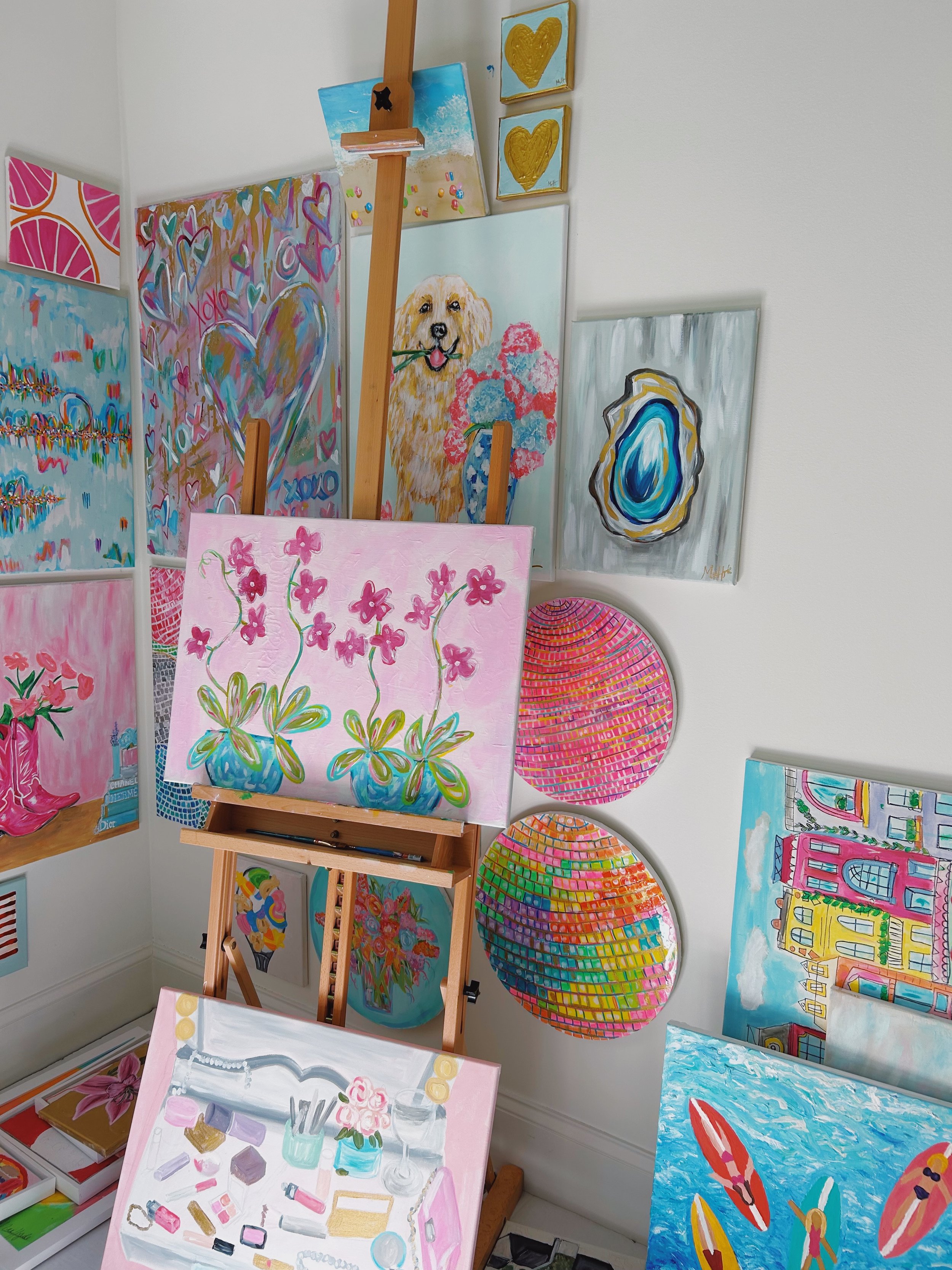 A collection of colorful paintings displayed on walls and an easel, including abstract designs, hearts, a dog, shells, and floral artwork, with some paintings leaning against the wall and others hanging.