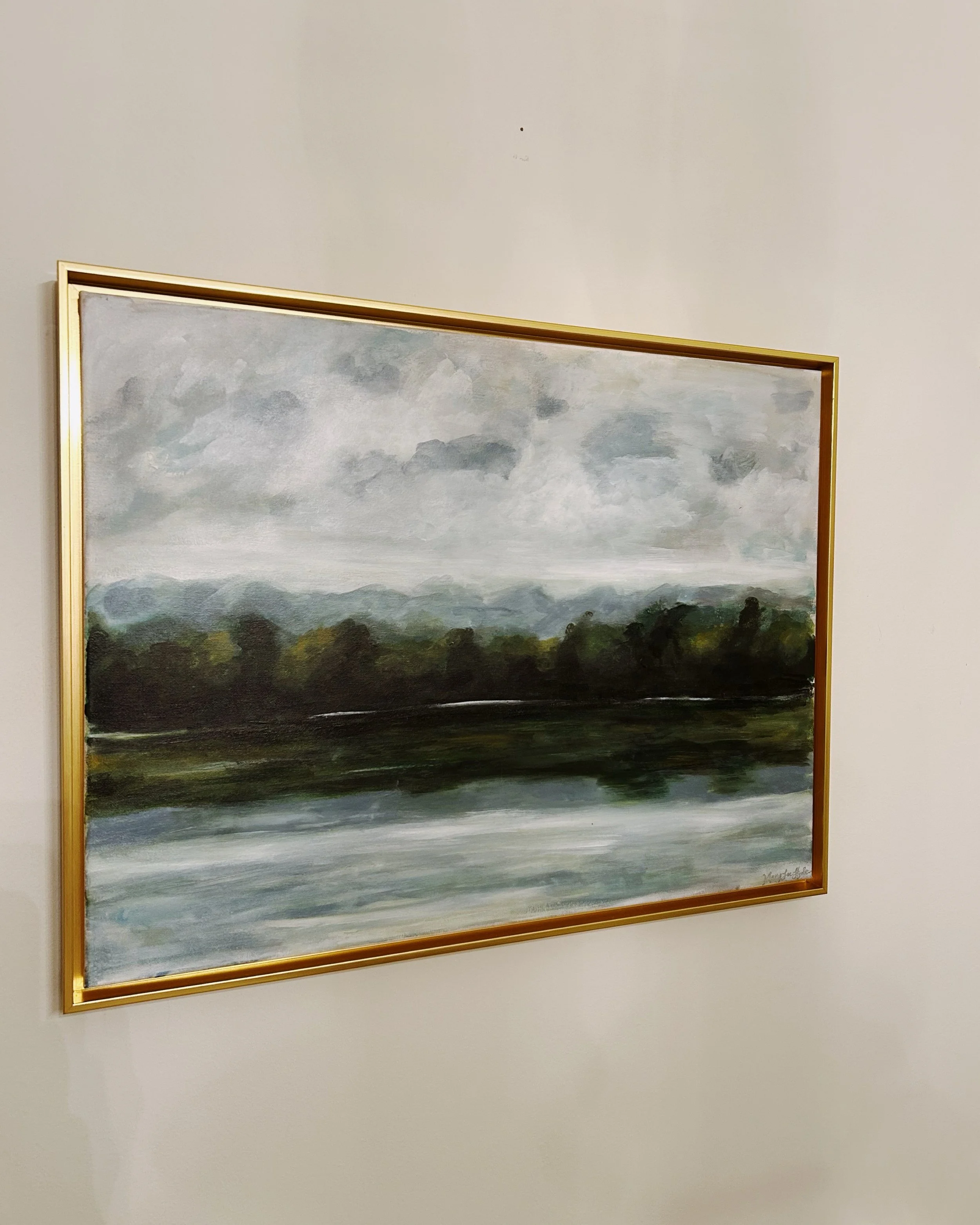 Landscape painting of trees and water under a cloudy sky, framed in gold, hanging on a plain white wall.