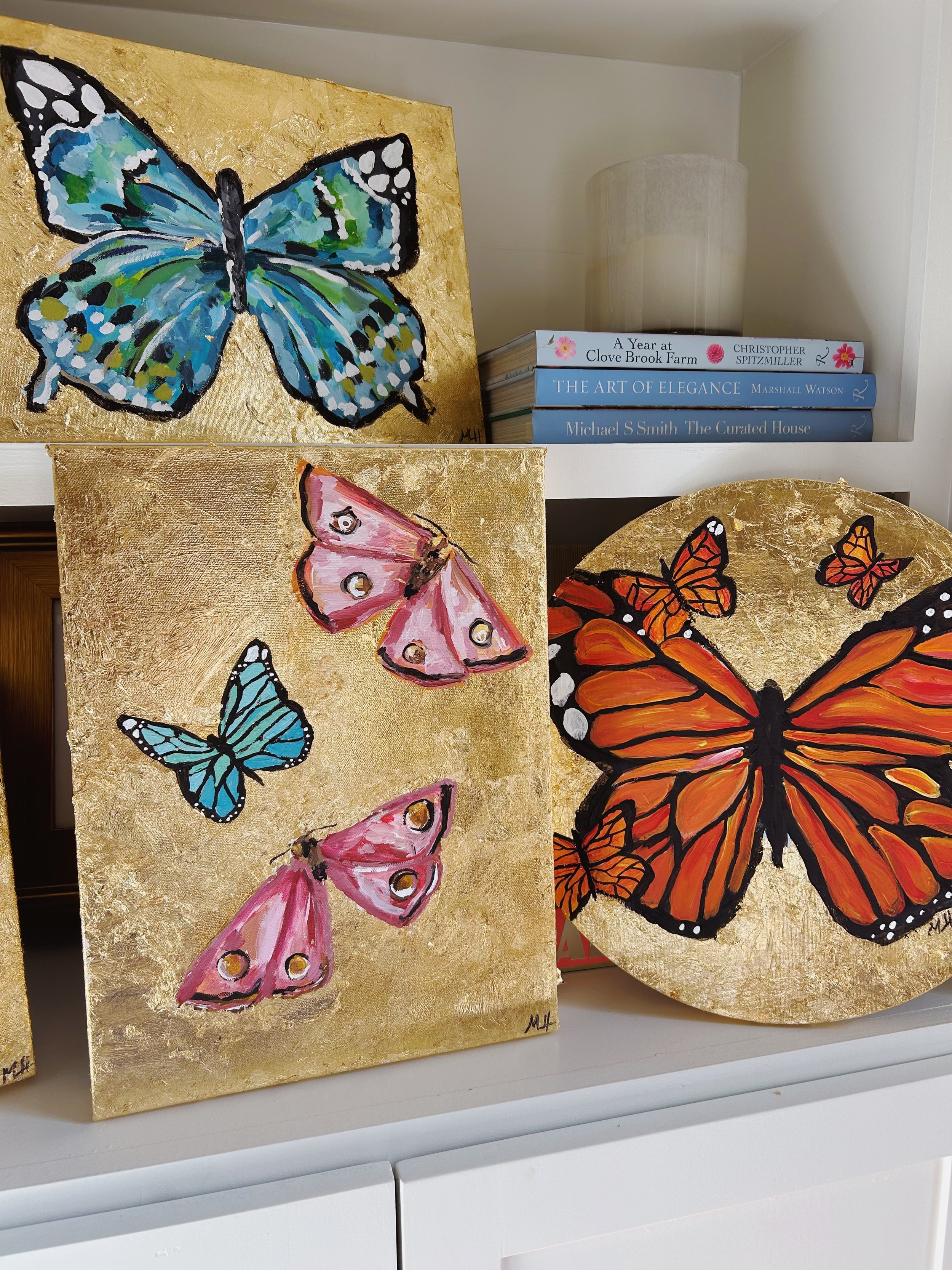 Three butterfly paintings with gold backgrounds displayed on a shelf; two rectangular paintings and one round painting, each featuring colorful butterflies.