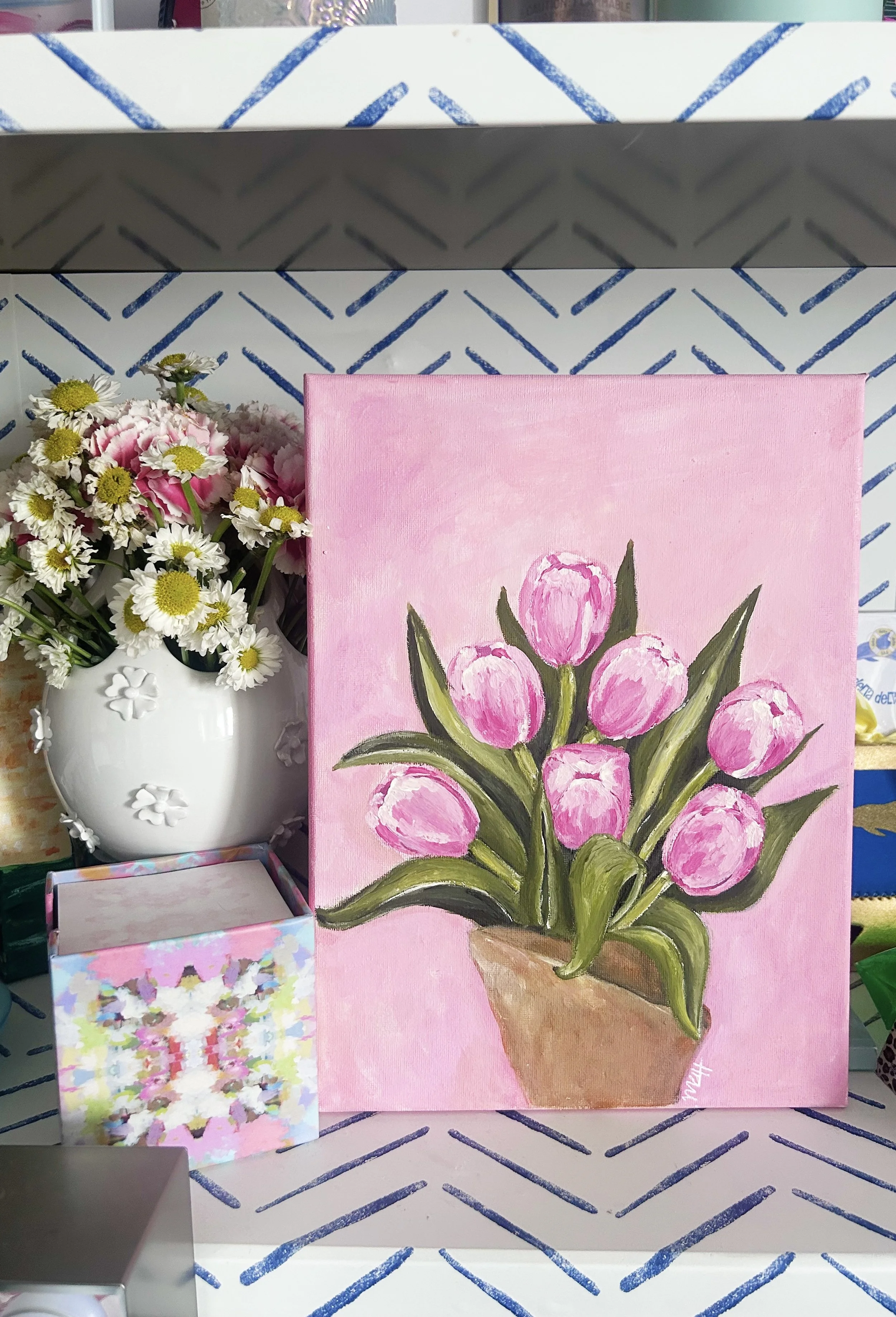 A pink canvas painting of a bouquet of pink tulips in a brown pot, placed on a shelf with a white vase of daisies and pink flowers to the left.