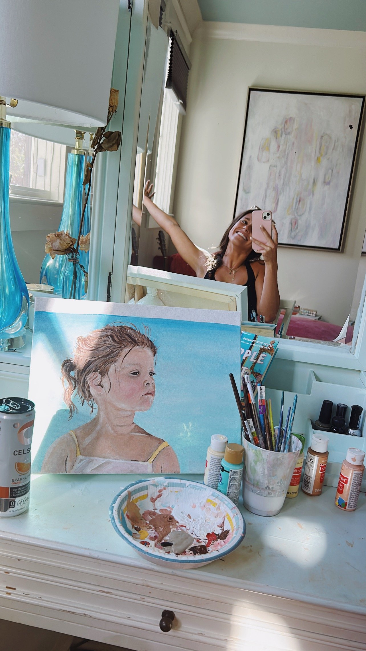 A woman taking a selfie in a mirror, smiling, with a painter's workspace in front of her. The workspace has a partially completed portrait of a young girl on an easel, several paint bottles, brushes in a cup, a plate with mixed paint, and a container