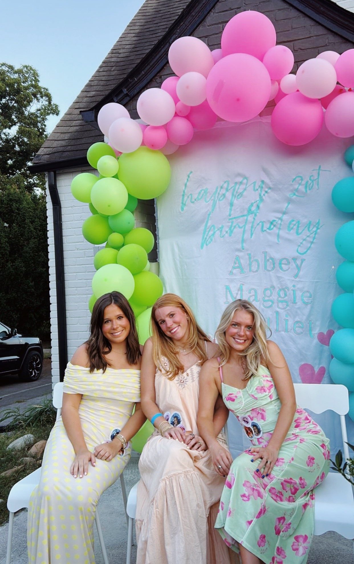 Three women sitting on white chairs at a birthday celebration outdoors, with a backdrop of pink, white, green, and teal balloons and a banner reading 'happy 21st birthday Abbey Maggie Olliet.'