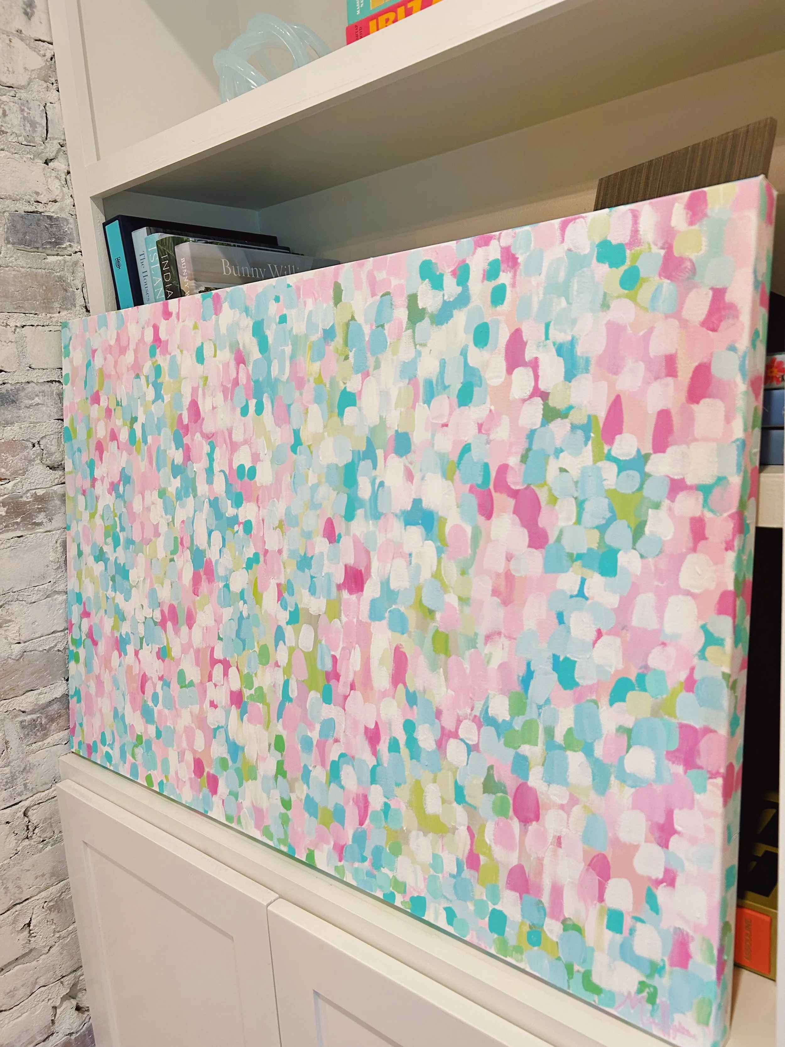A colorful abstract painting with small, rounded patches of pink, blue, white, and green, displayed on a shelf or cabinet.