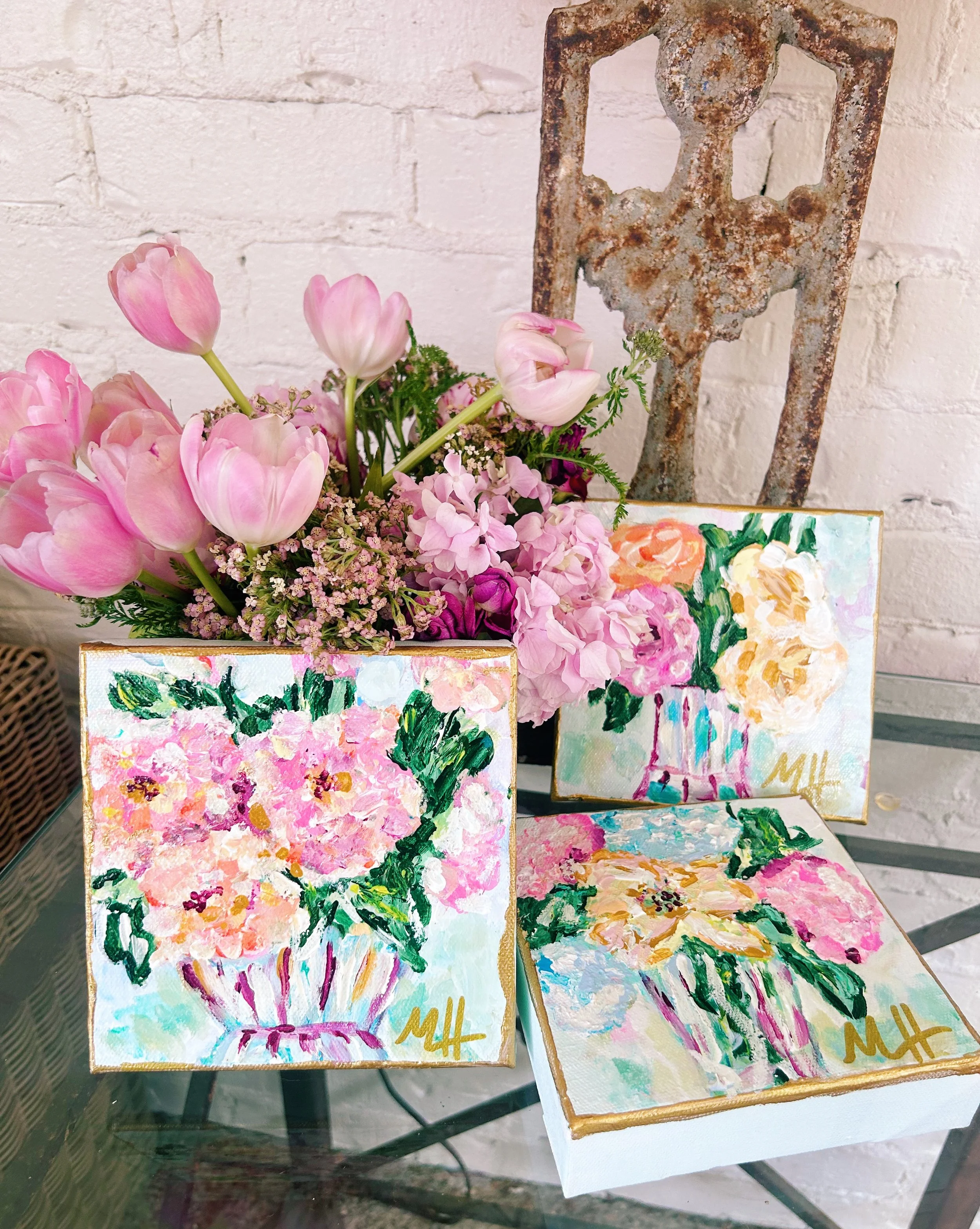A bouquet of pink tulips and purple flowers in a vase with three small floral paintings on a glass table, against a white brick wall.