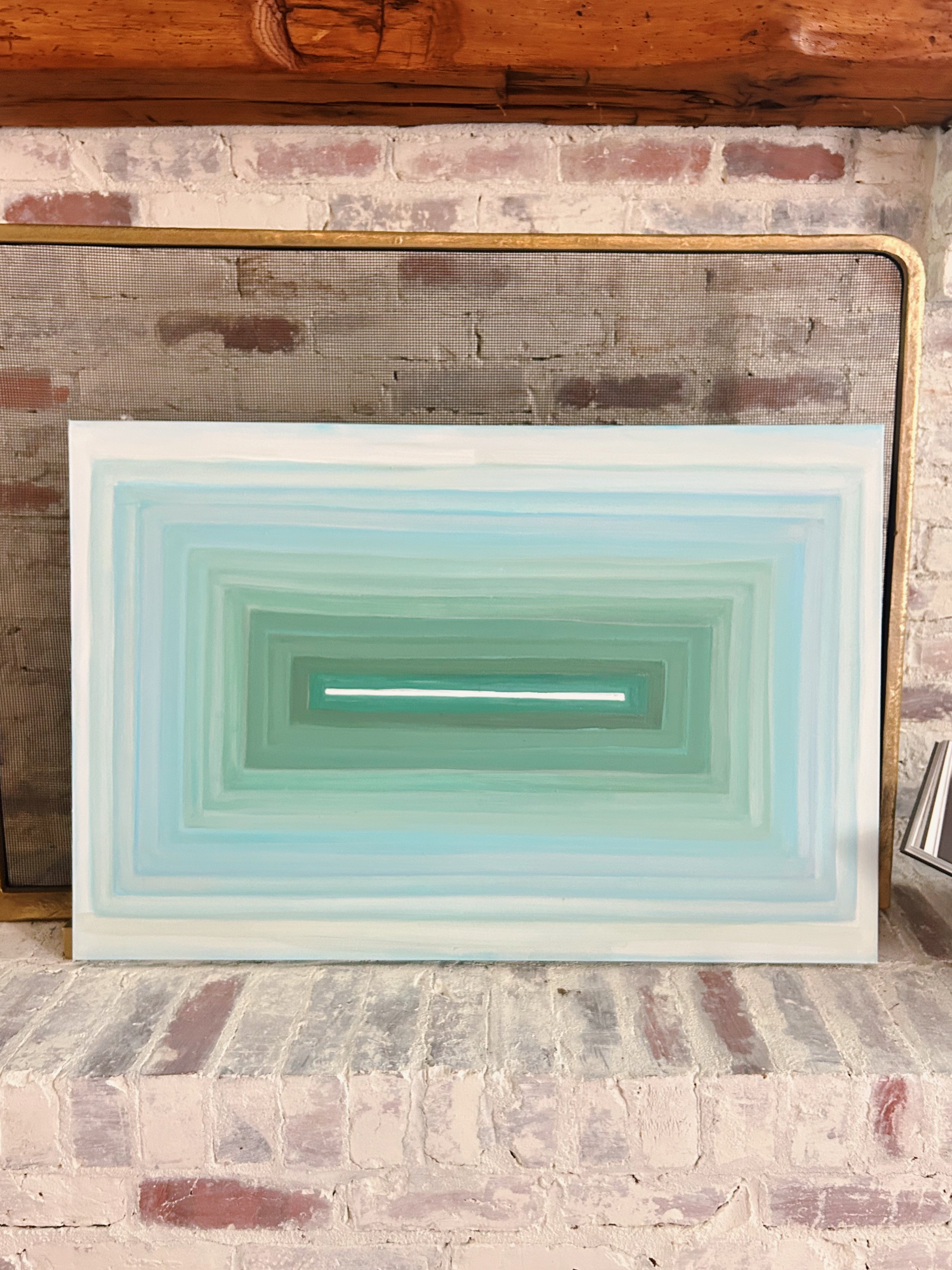 A painting with concentric rectangles in shades of green and blue, resting on a brick hearth in front of a brick wall and fireplace.