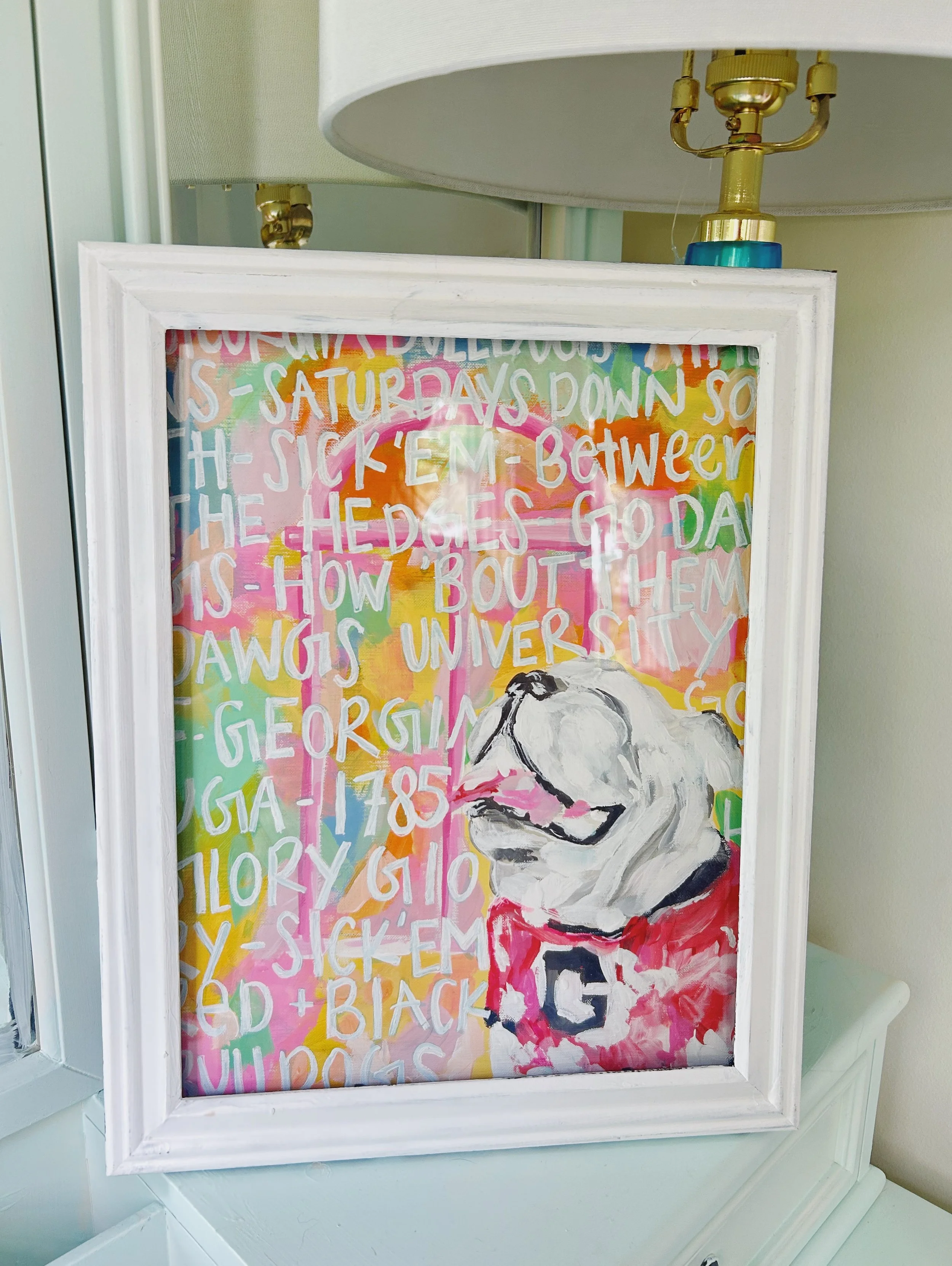 Colorful painting of a bulldog with a pink background, white and yellow text about Bulldogs, and a fluffy white bulldog wearing a red shirt.