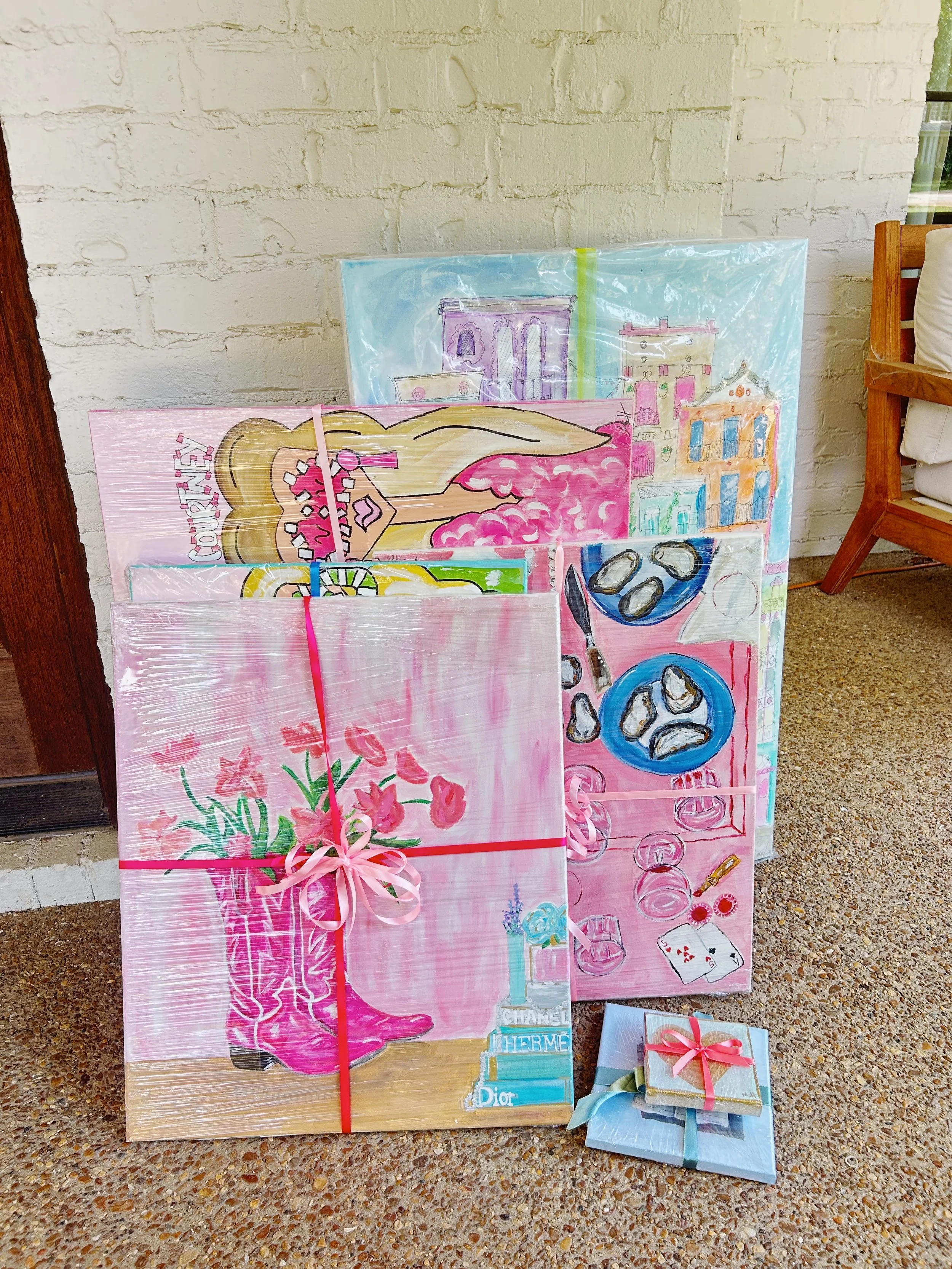 Collection of colorful artwork and wrapped gift boxes leaning against a white brick wall. The artwork features images of buildings, oysters, a girl with blonde hair, pink boots, and books with brand labels like Chanel, Hermès, and Dior.