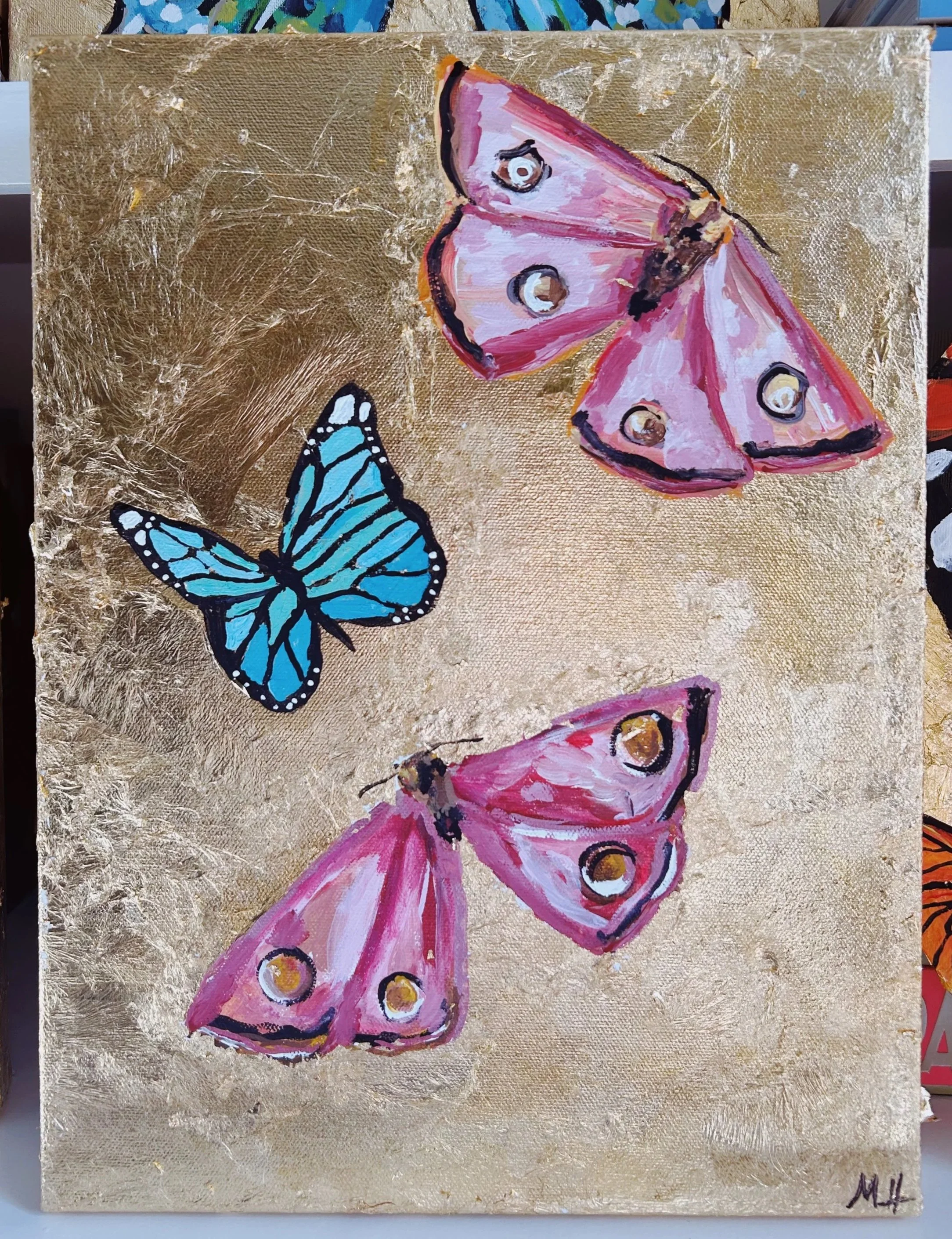 Painting of two pink moths with yellow and black eyespots on their wings and a blue butterfly with black and white wing patterns, set against a textured gold background.