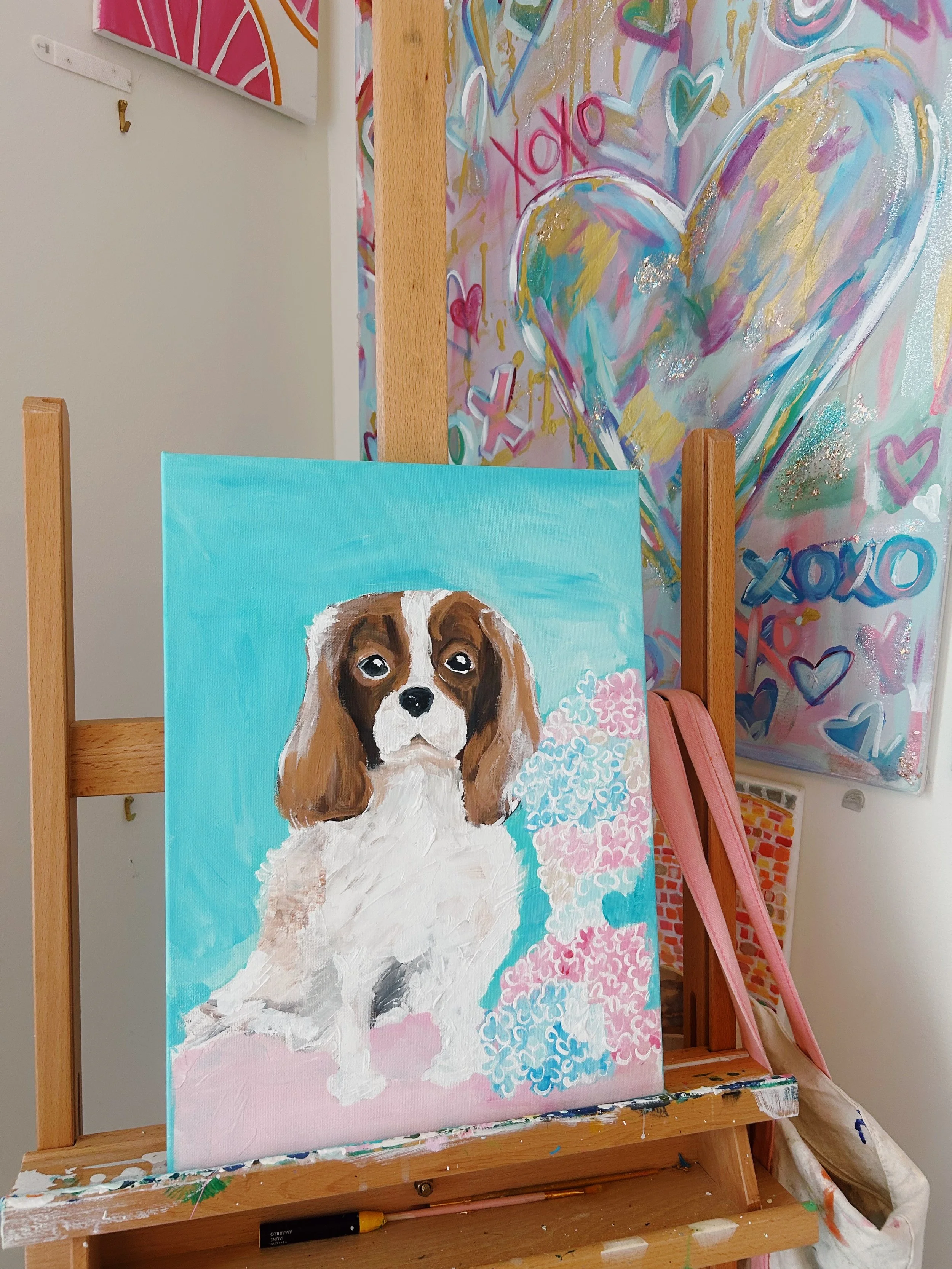 Painting of a brown and white dog with sad eyes on a blue background, surrounded by pink and white flowers, on an easel in an art studio.