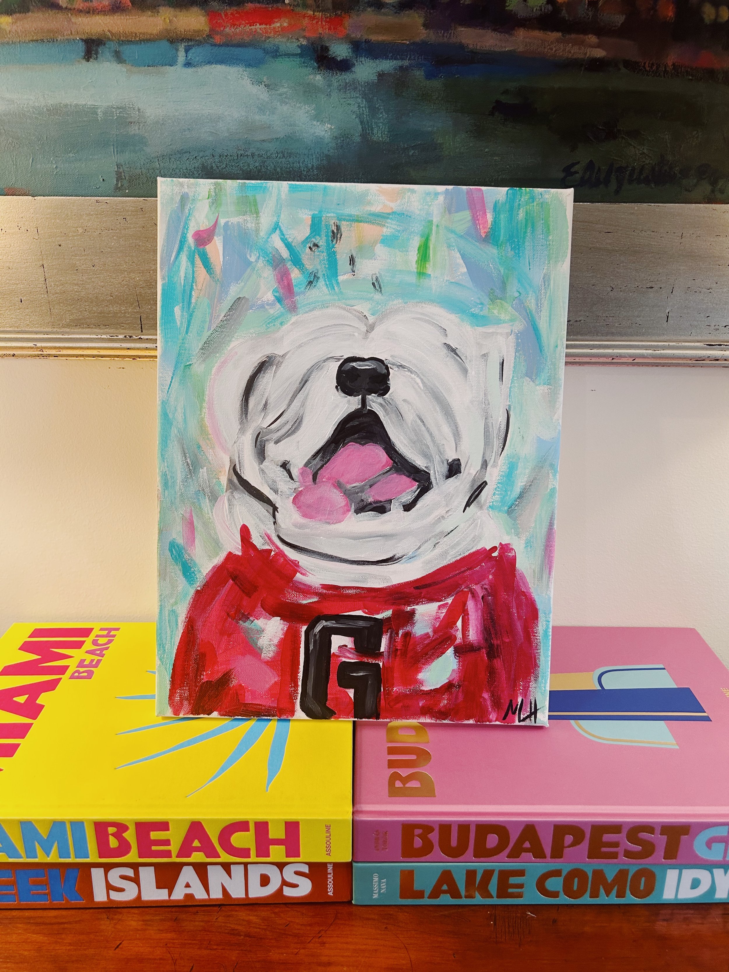Painting of a white dog with a black nose, pink tongue, and red hoodie, on top of colorful books about Miami Beach, Budapest, and Lake Como.