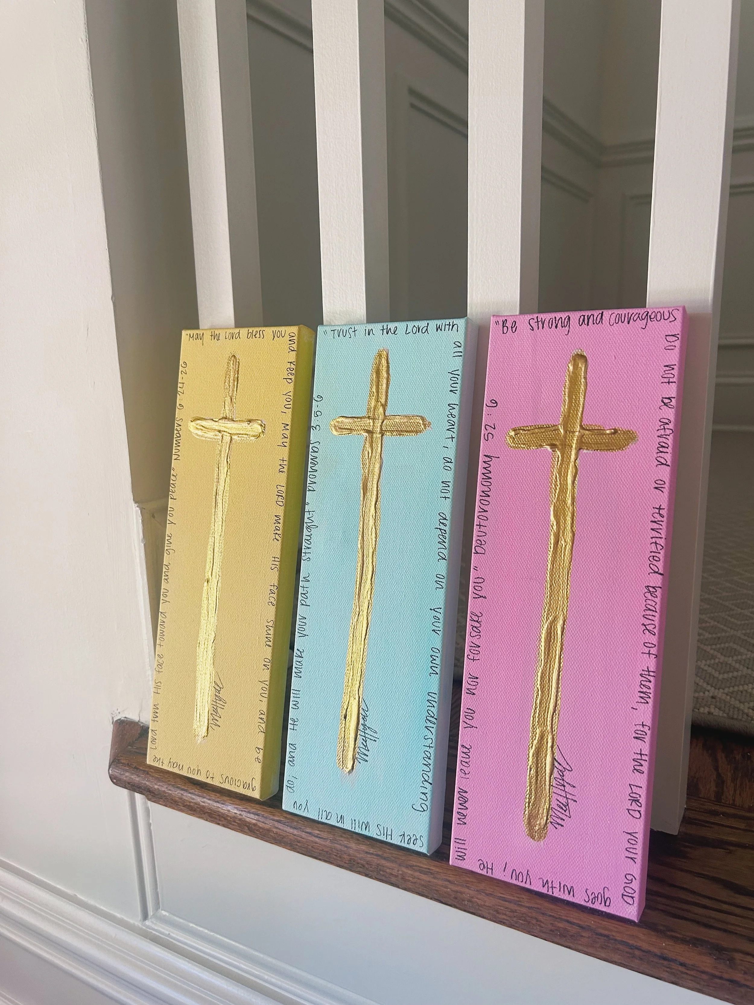 Three decorated canvases with gold crosses on pastel backgrounds of yellow, blue, and pink, each with handwritten Bible verse around the edges.
