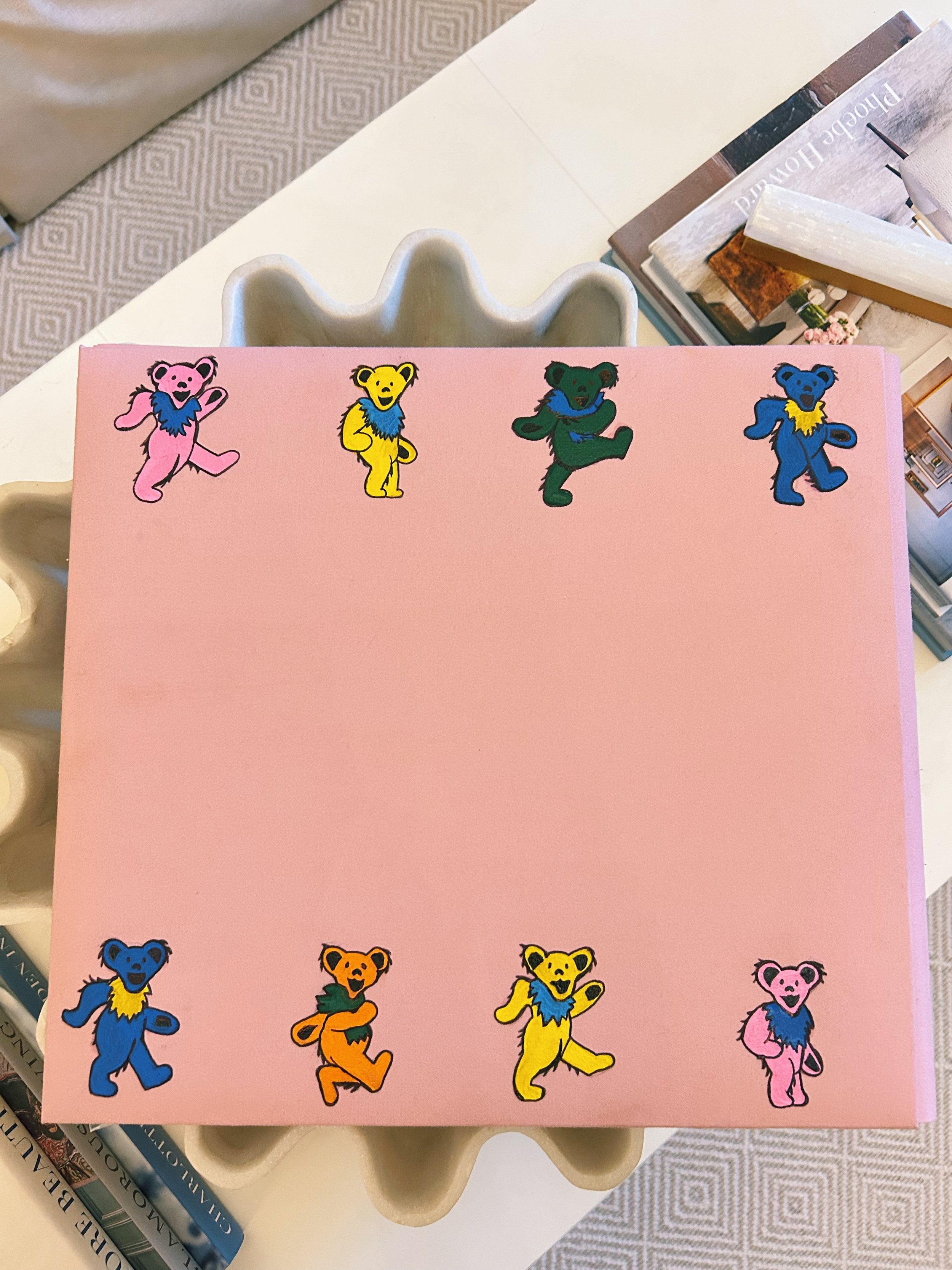 Pink box with colorful teddy bear stickers arranged around the edges, set on a table with magazines and decorative objects.