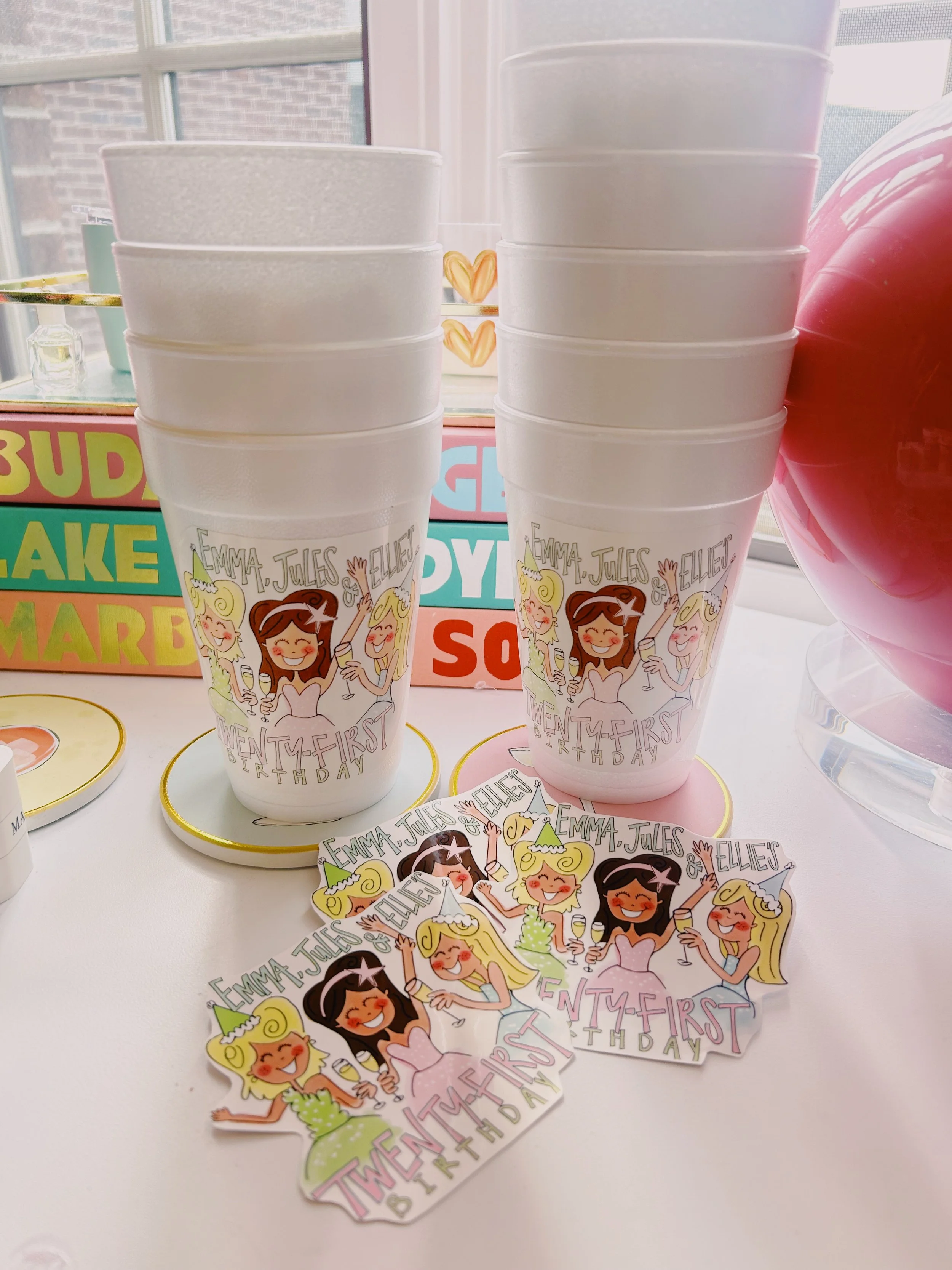 Stacked white paper cups with birthday-themed illustrations of girls celebrating, labeled with the names Emma, Jules, and Ellie, and the phrase 'Twenty-First Birthday'.