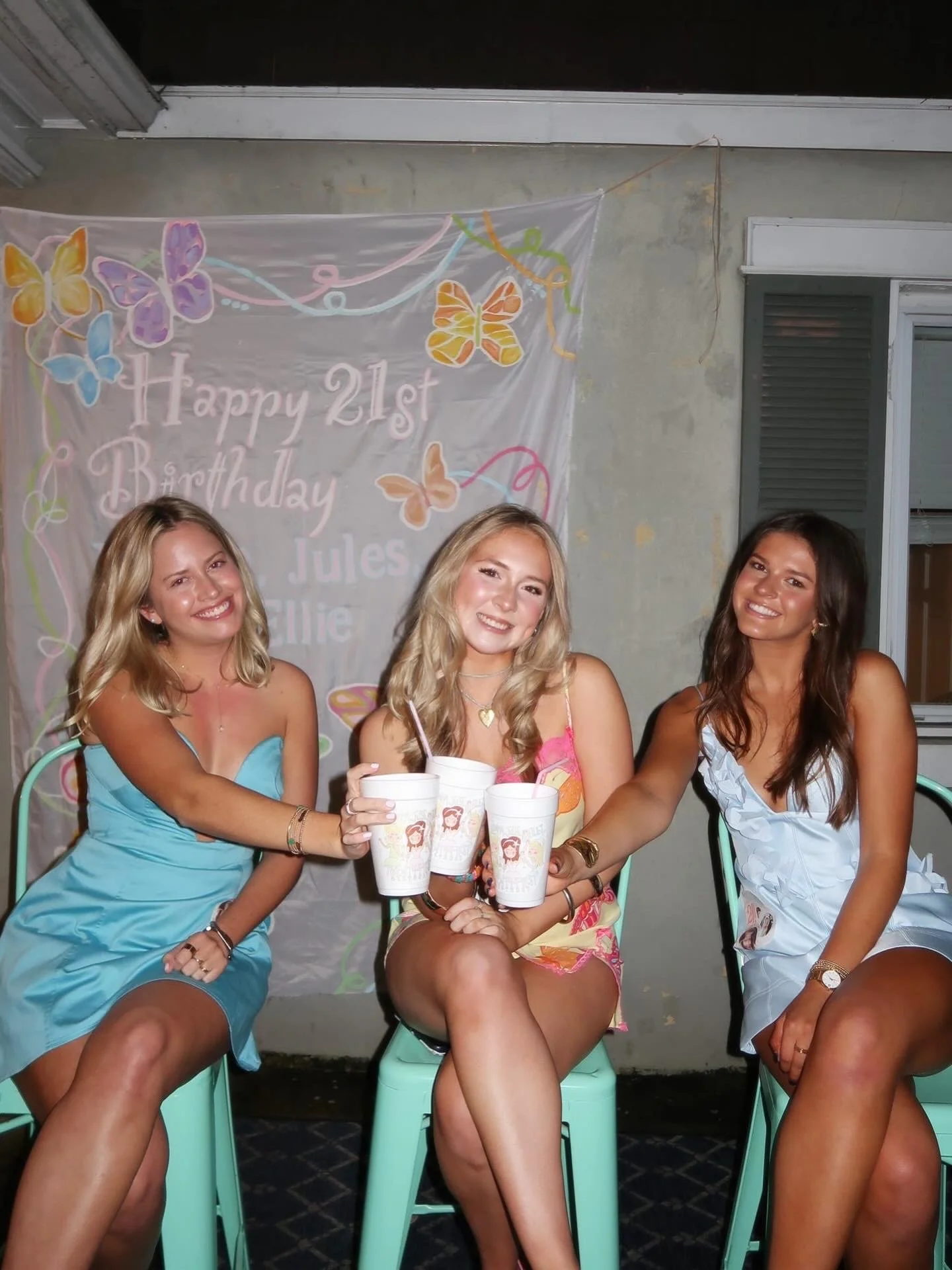 Three women sitting and smiling at a birthday celebration in front of a colorful butterfly-themed banner that reads 'Happy 21st Birthday Jules Ellie'. They are holding drinks and appear to be enjoying the party.