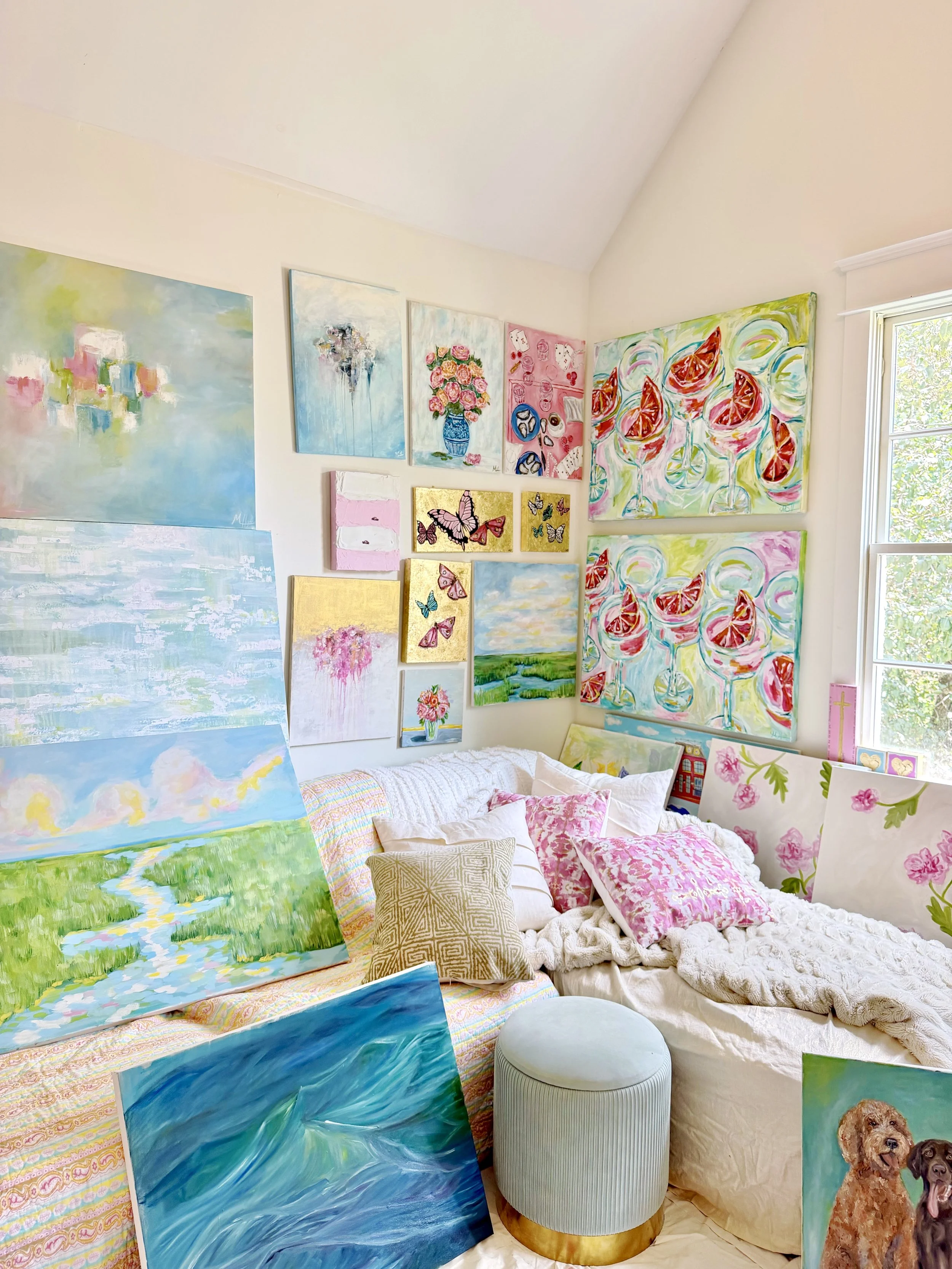 A cozy room filled with colorful paintings on the walls and propped against furniture, featuring abstract landscapes, floral arrangements, butterflies, and a table with pillows, a small stool, and a window with sunlight.