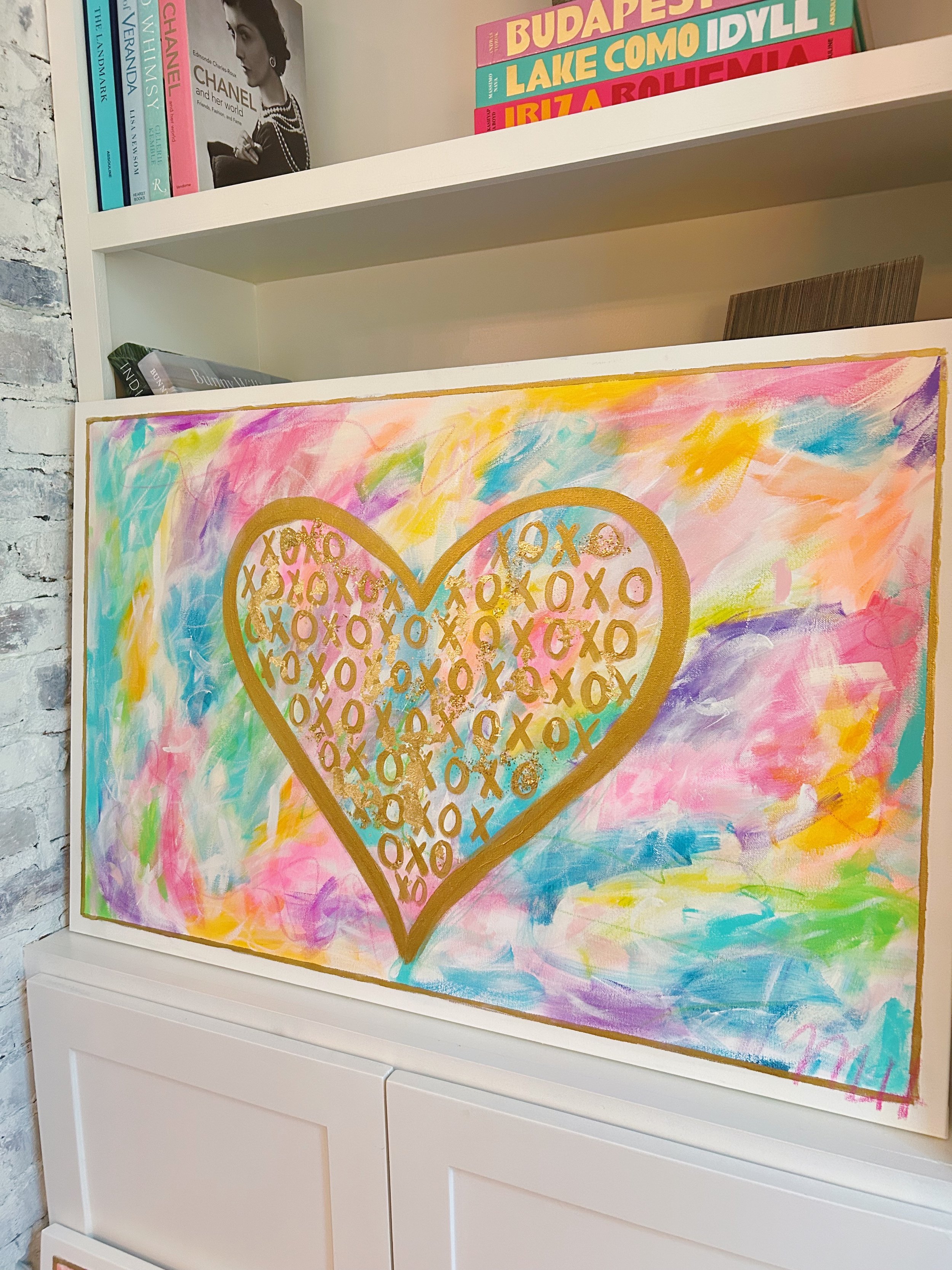Colorful abstract painting of a gold-outlined heart filled with gold and pink 'X' and 'O' symbols, with a multicolored background of pink, blue, yellow, and purple brushstrokes, placed on a white shelf.