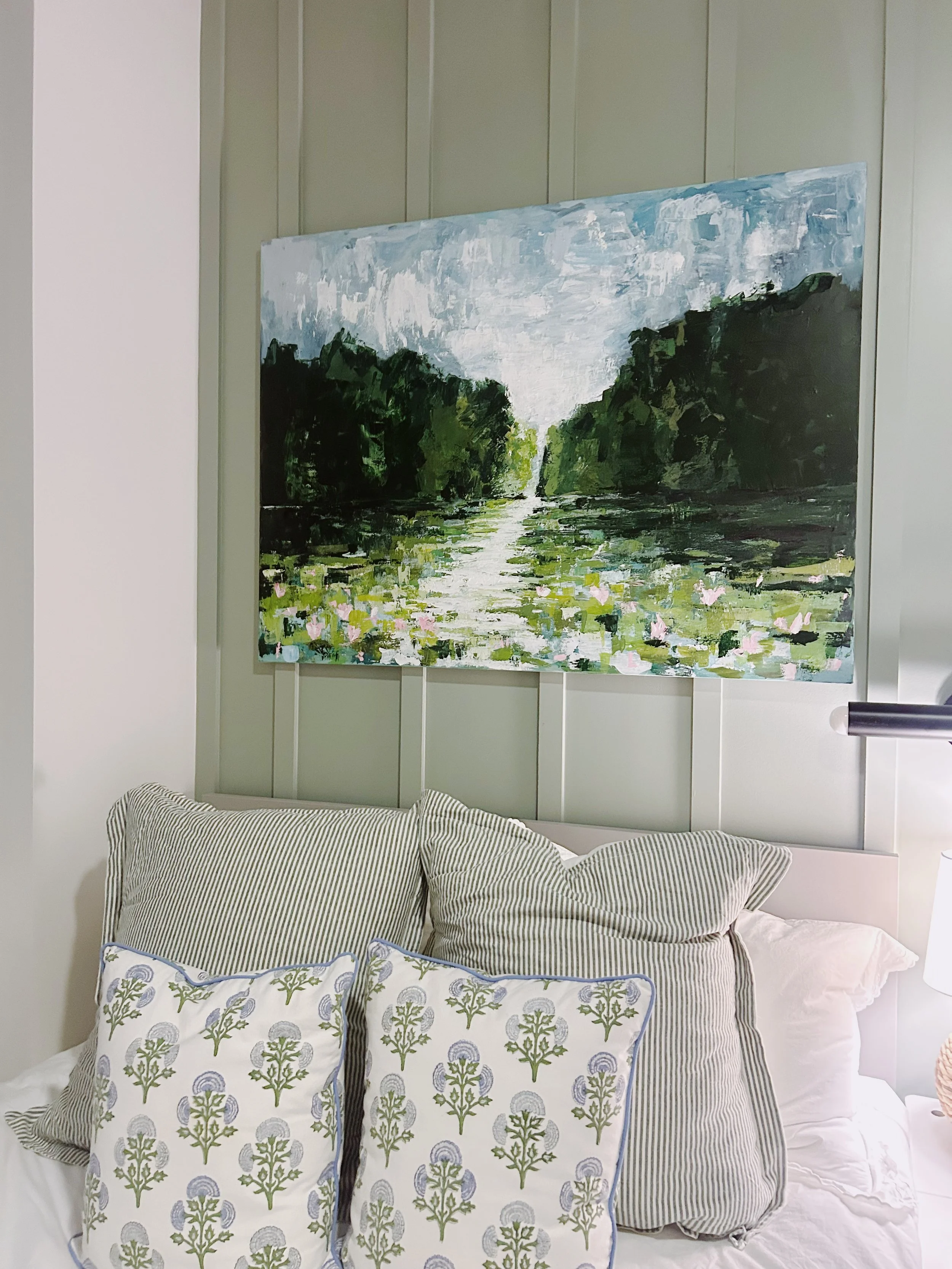 A bedroom with a landscape painting of a river with greenery and water lilies hanging above the bed. The bed has pillows with green and white floral and striped patterns.