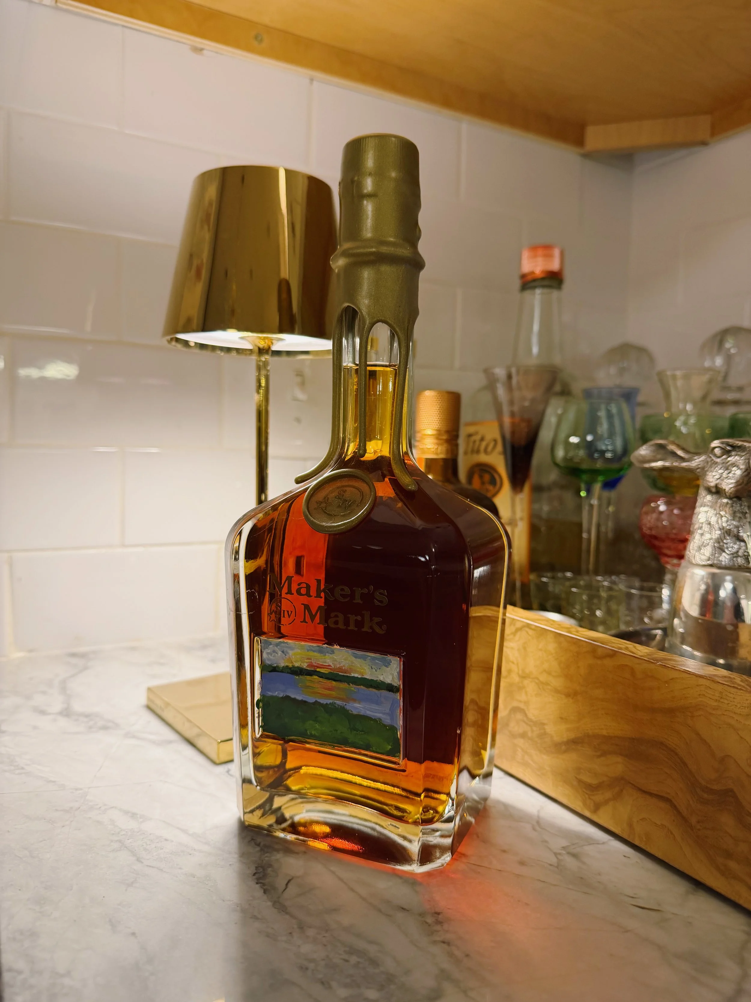 A bottle of Maker's Mark bourbon whiskey with a gold foil seal on top, placed on a marble countertop. Behind the bottle, there is a gold-colored table lamp, a wooden tray, a variety of glassware, and liquor bottles on a shelf with a white tiled wall 
