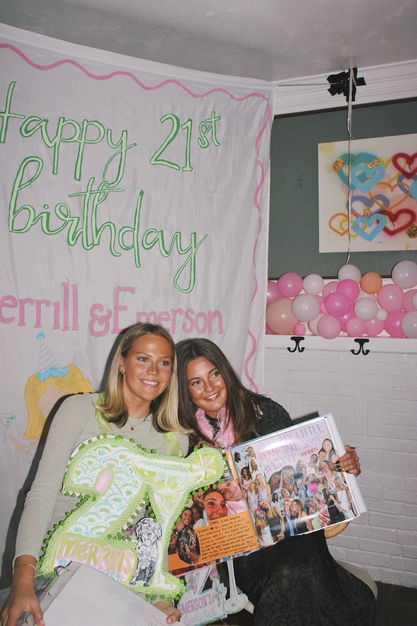 Two women celebrating a 21st birthday at a party, holding a scrapbook with photos and decorations, with a banner reading 'happy 21st birthday Merrill & Emerson' behind them, and pink and white balloons in the background.