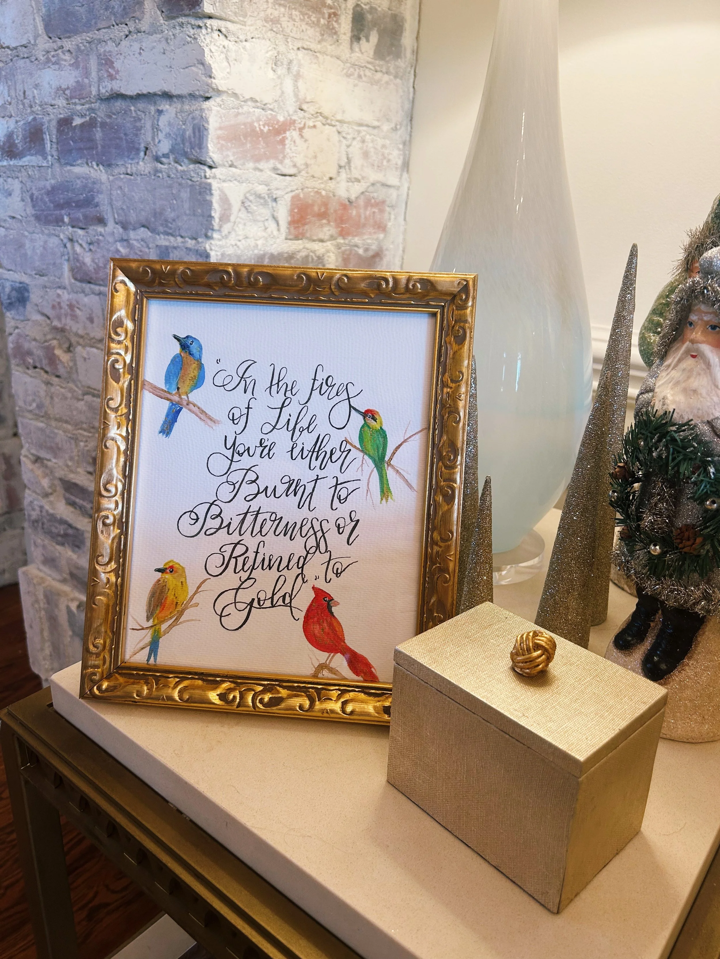 Decorative framed quote surrounded by colorful bird illustrations on a table with Christmas ornaments and a large white vase in the background.