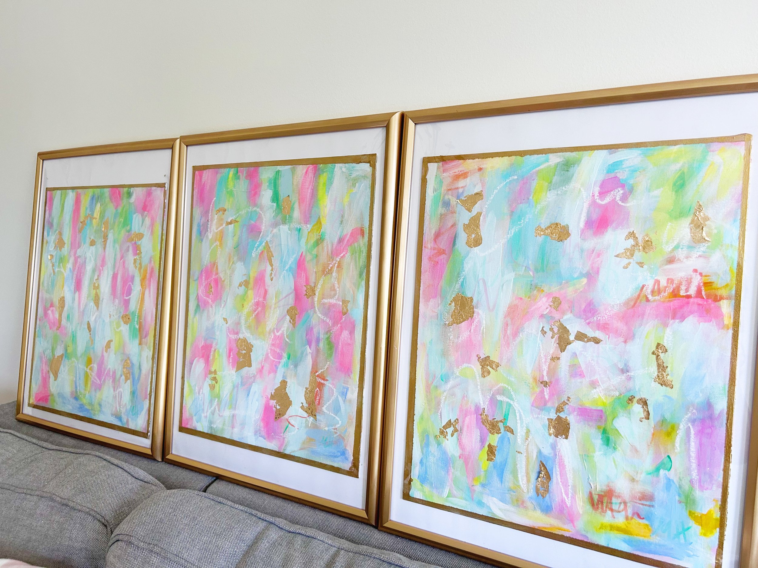 Three abstract paintings with pastel pink, blue, green, and white colors, accented with gold foil, framed in gold and resting against a white wall on a grey couch.