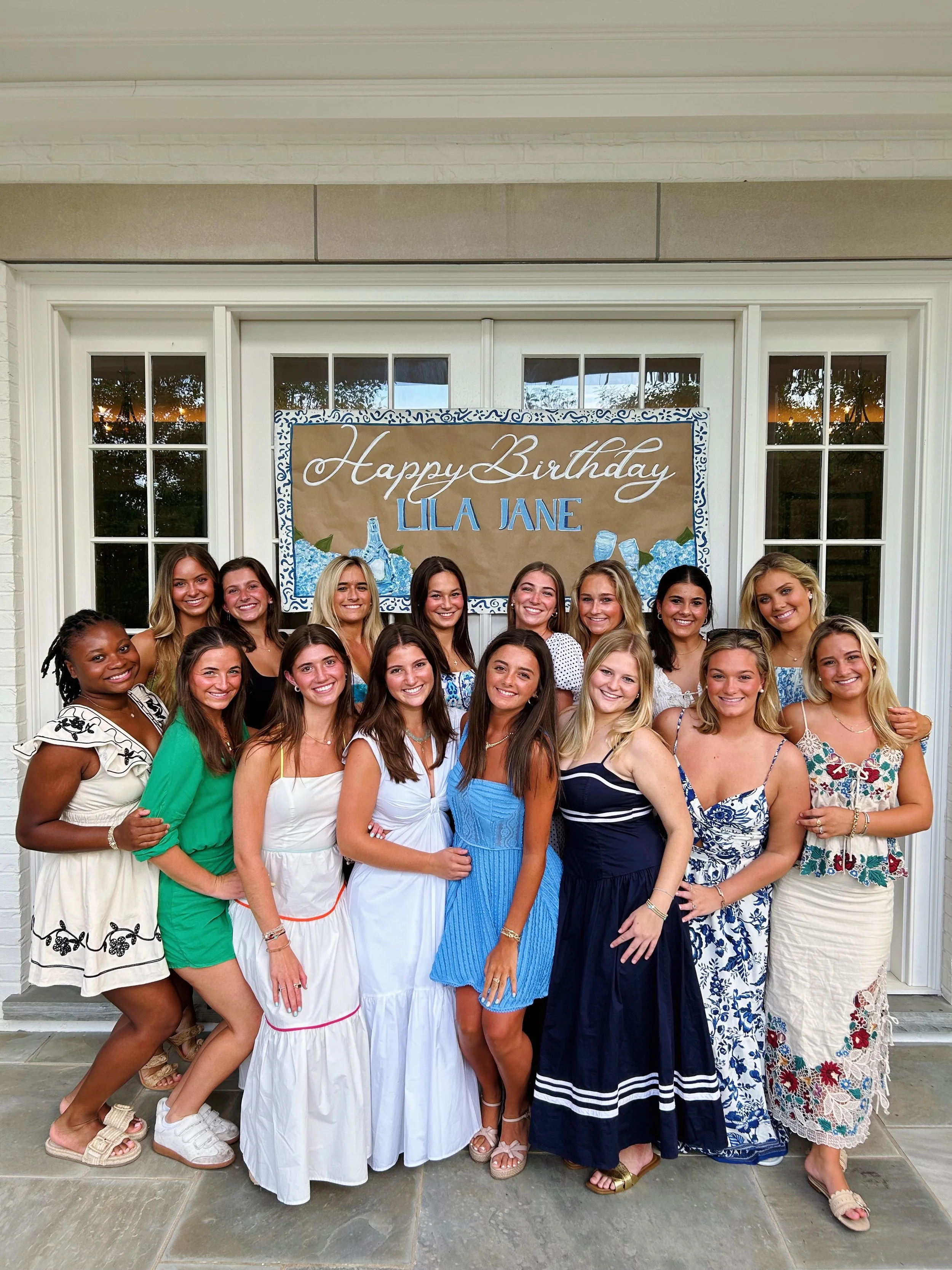 Group of young women posing together at a birthday party, standing in front of a large decorated sign that reads 'Happy Birthday Lila Jane', with windows and a door behind them.