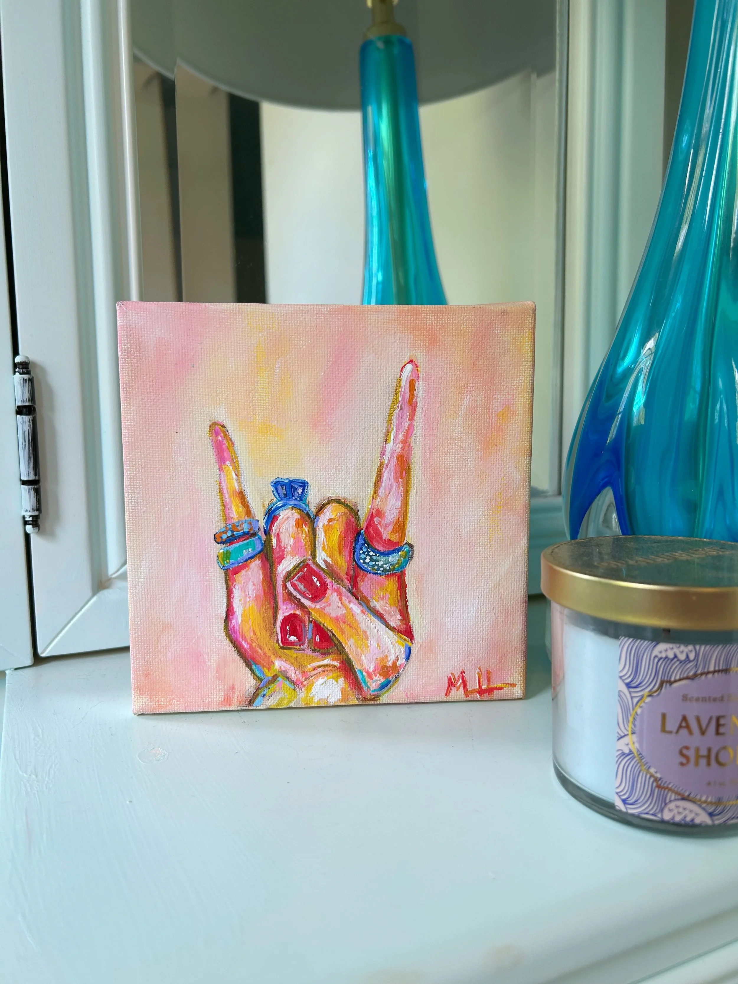 A small painting of a hand making the rock and roll gesture, with colorful rings on the fingers, signed with initials 'ML', placed on a white surface next to a scented candle labeled 'Lavender Shop' and blue decorative glass vases in the background.